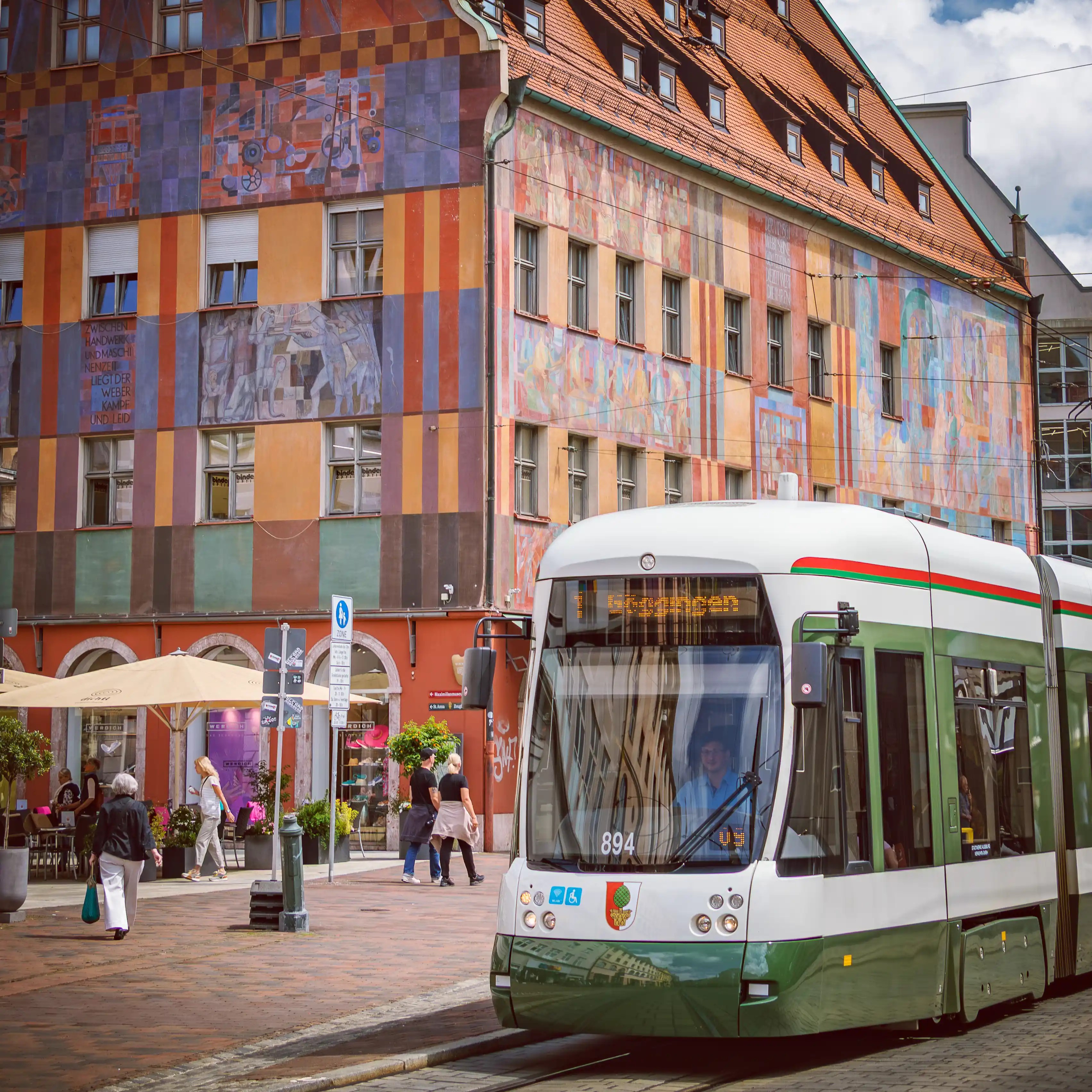 A modern tram passes a colorful mural-covered historic building in Augsburg’s city center.