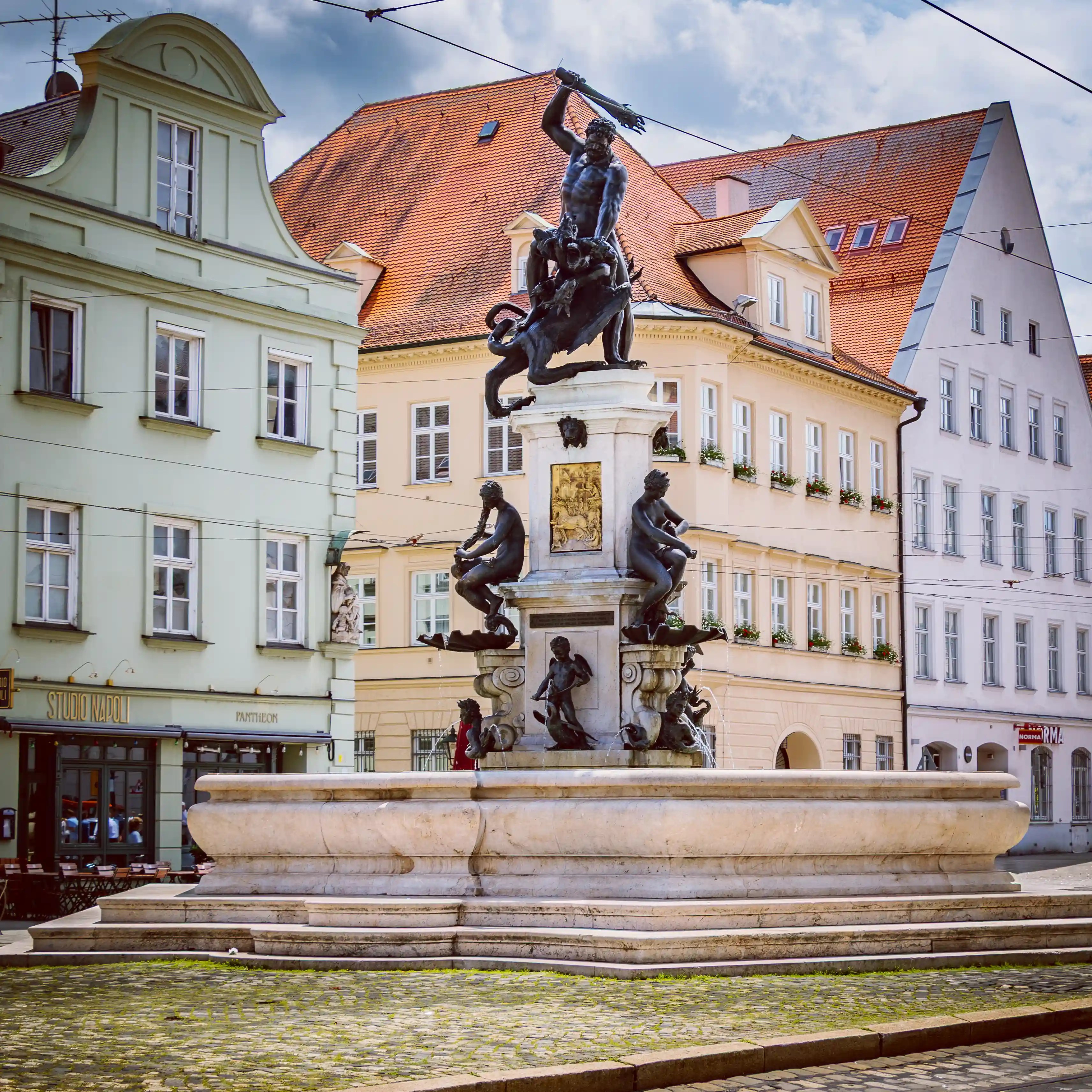 The Herkulesbrunnen fountain in Augsburg featuring a bronze statue of Hercules atop a sculpted pedestal surrounded by historic buildings.