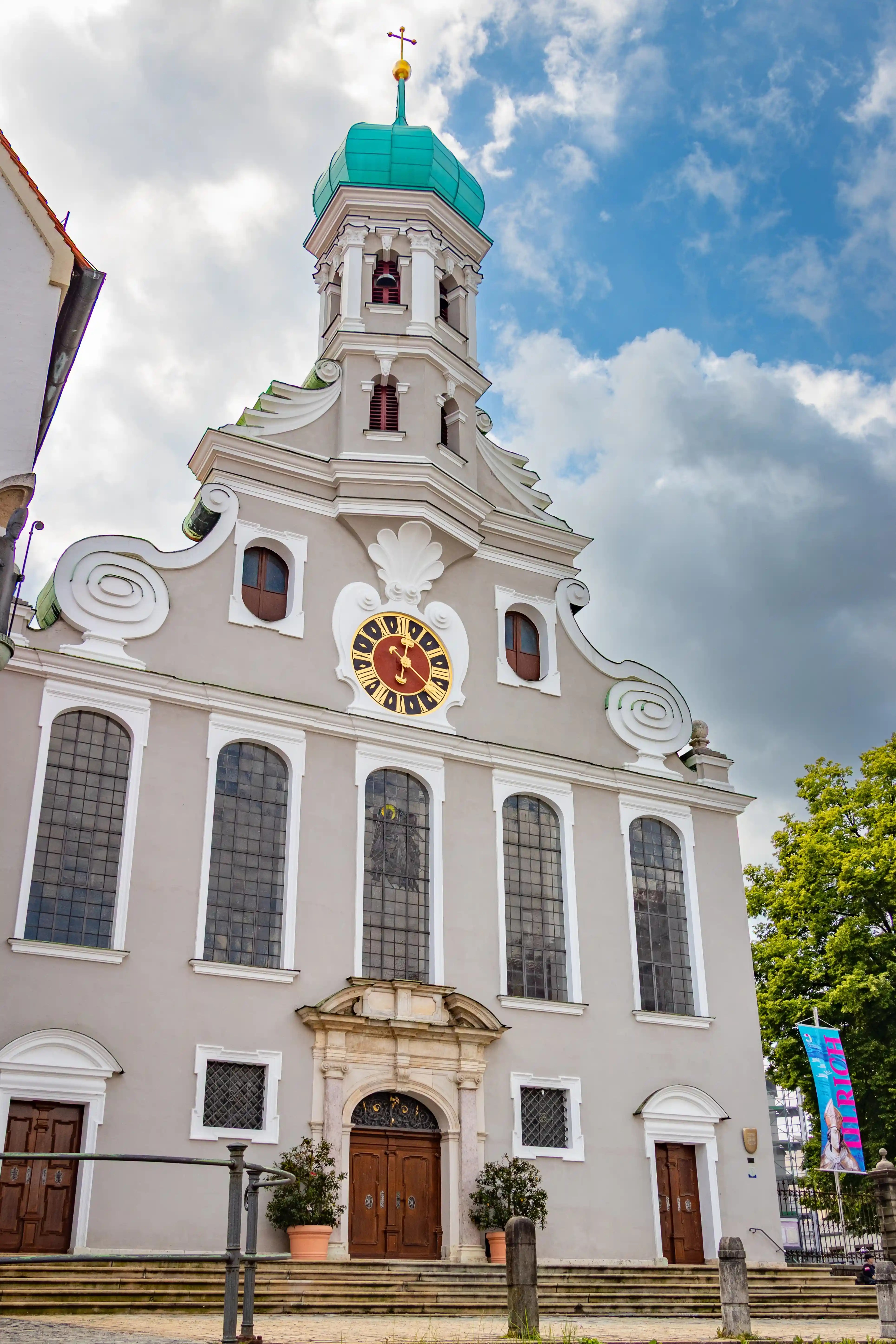 The façade of St. Ulrich and Afra Basilica in Augsburg with one visible tower topped by a turquoise dome and a large clock.