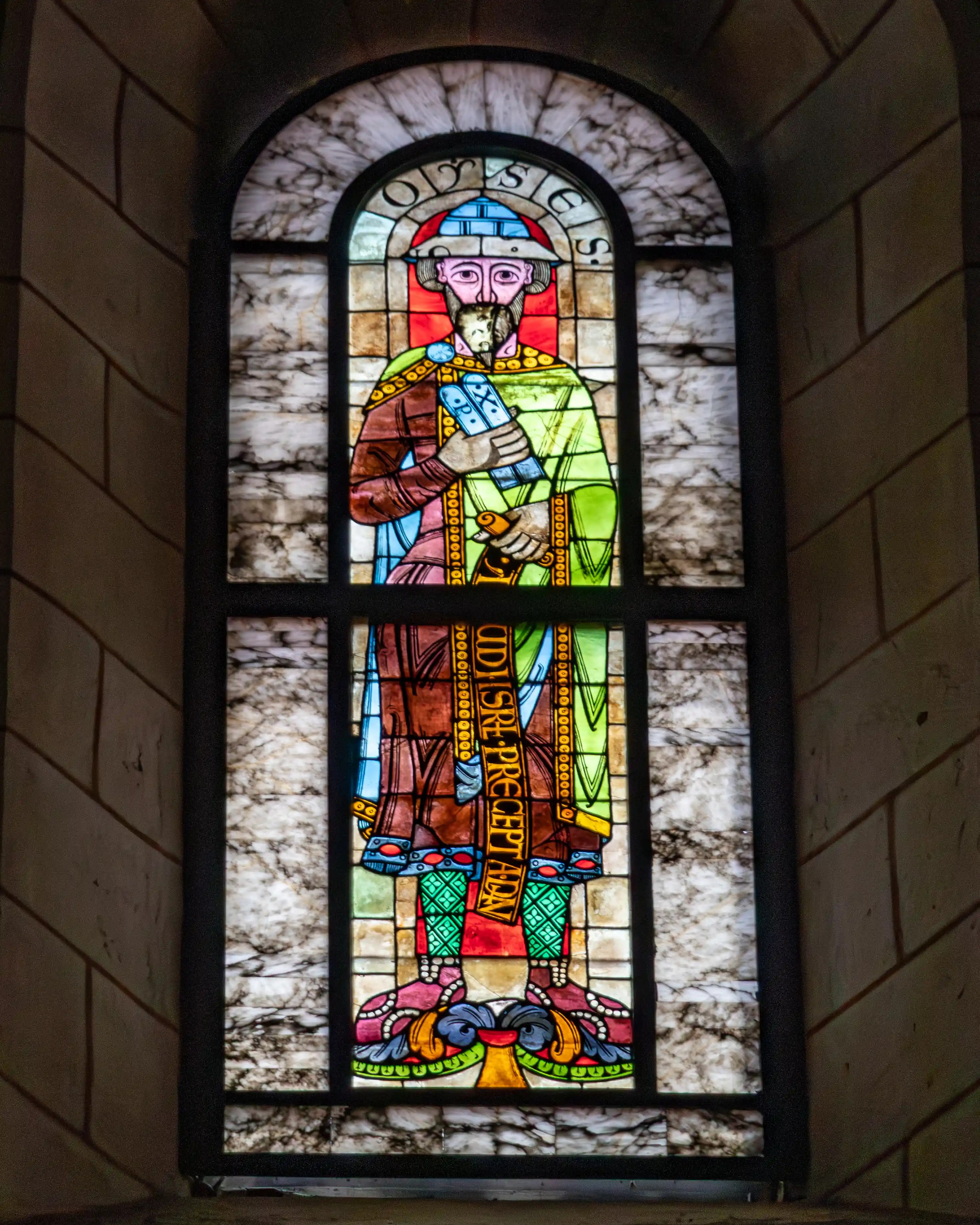 A stained glass window in Augsburg Cathedral showing a standing figure holding a scroll, surrounded by patterned glass.