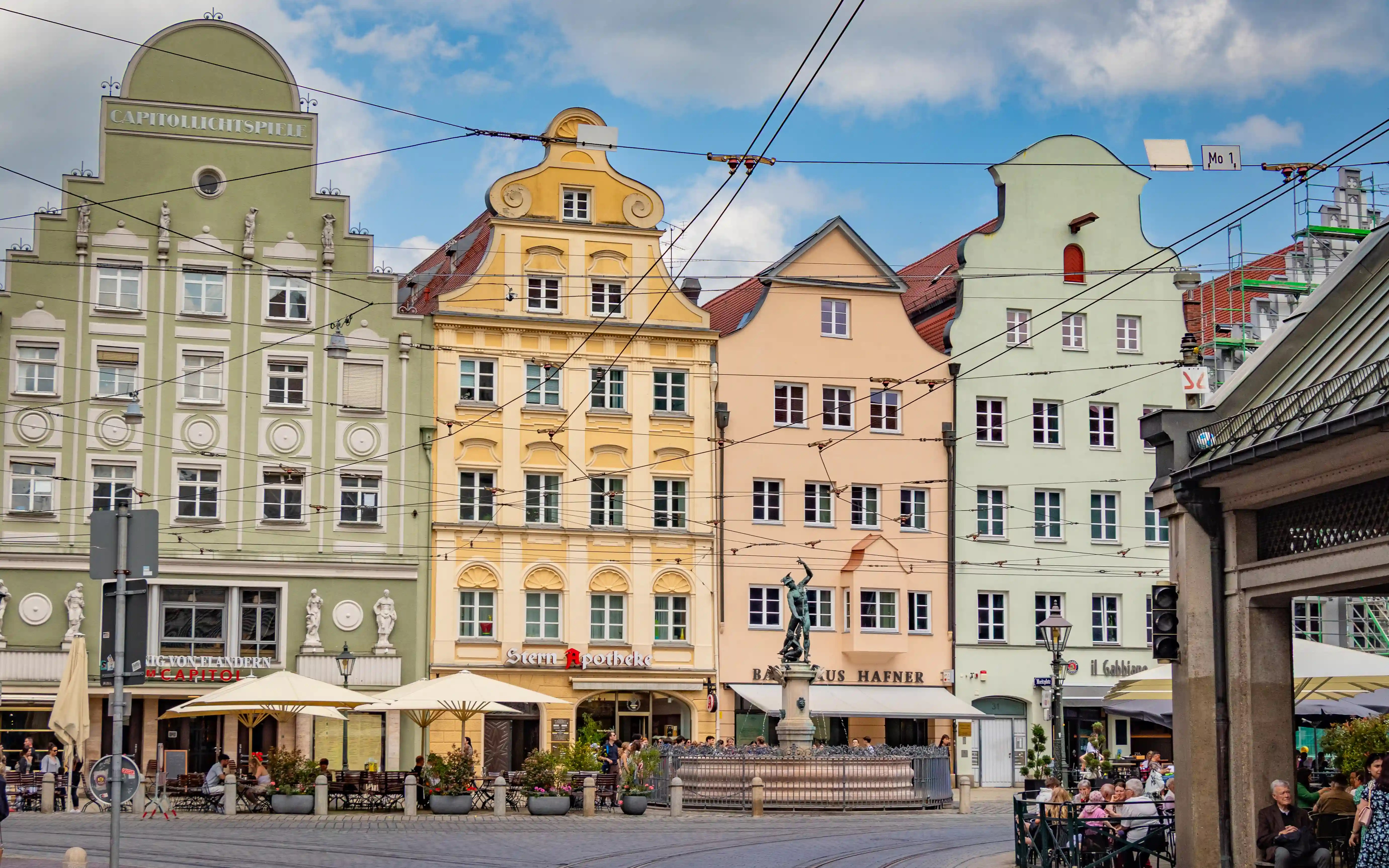 A city square in Augsburg lined with pastel-colored historic buildings, tram wires overhead, and a fountain at the center.