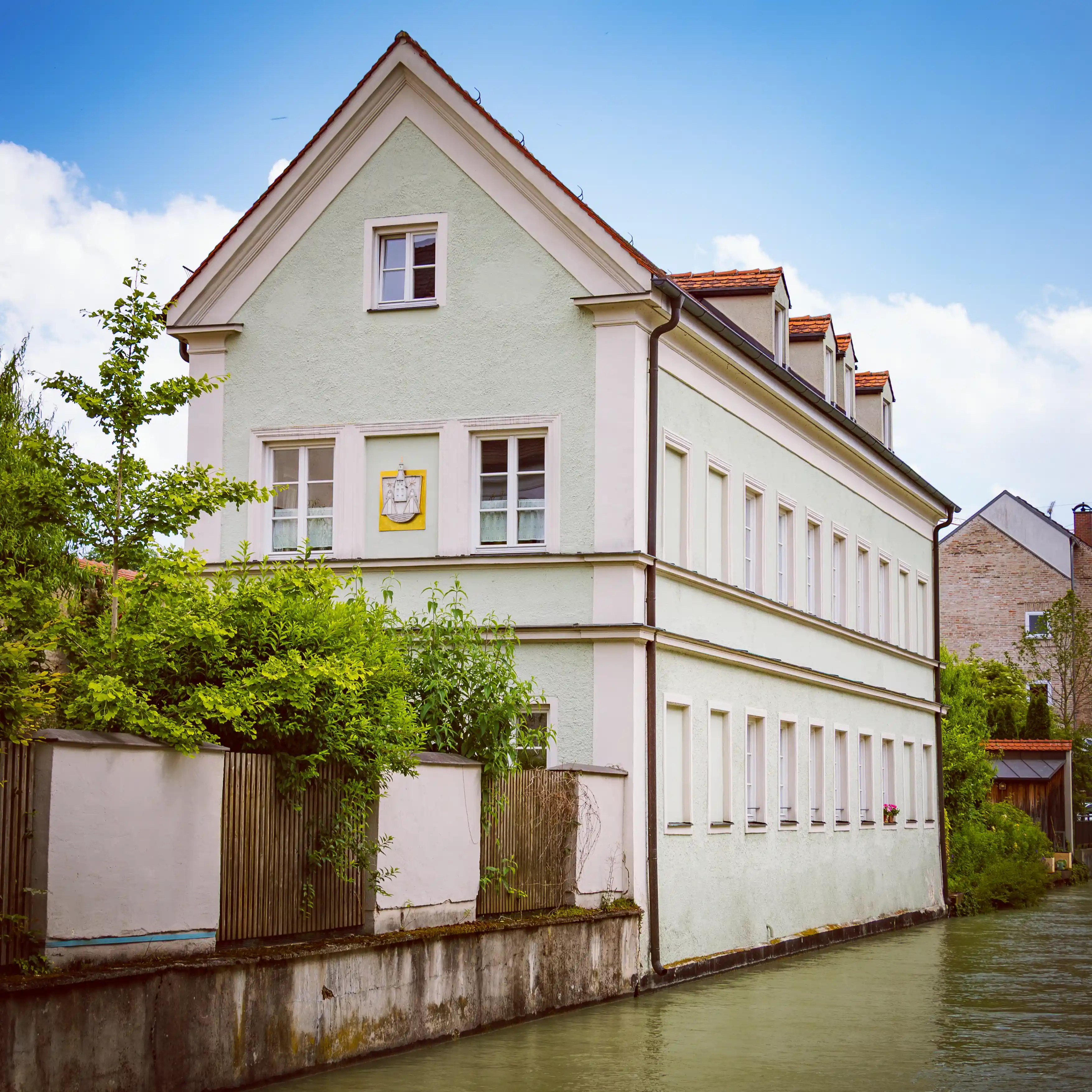 A pale green historic building in Augsburg standing directly beside a narrow canal with calm water.