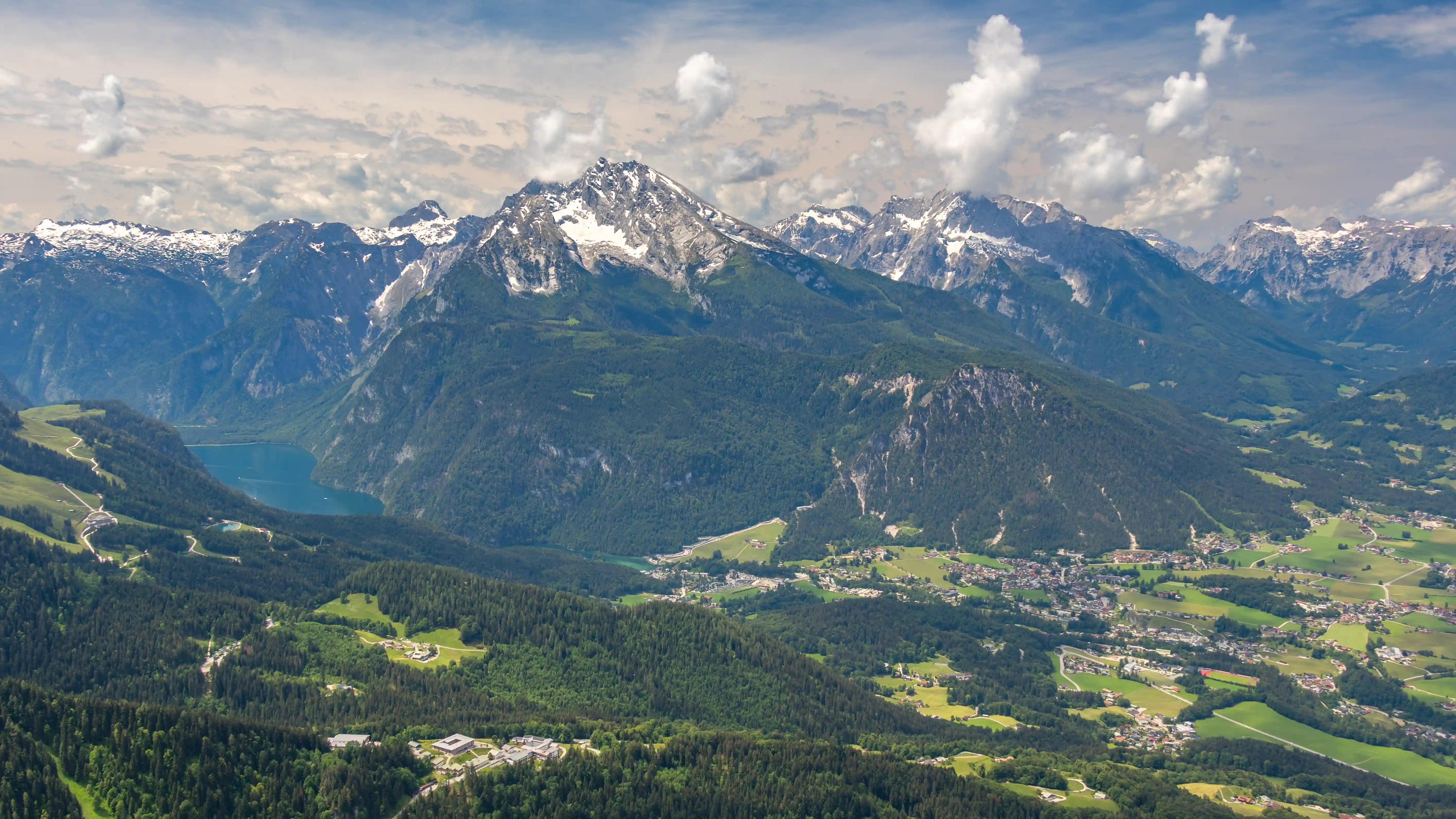 A wide alpine valley with scattered villages, green fields, and layered mountain ridges under a blue sky.