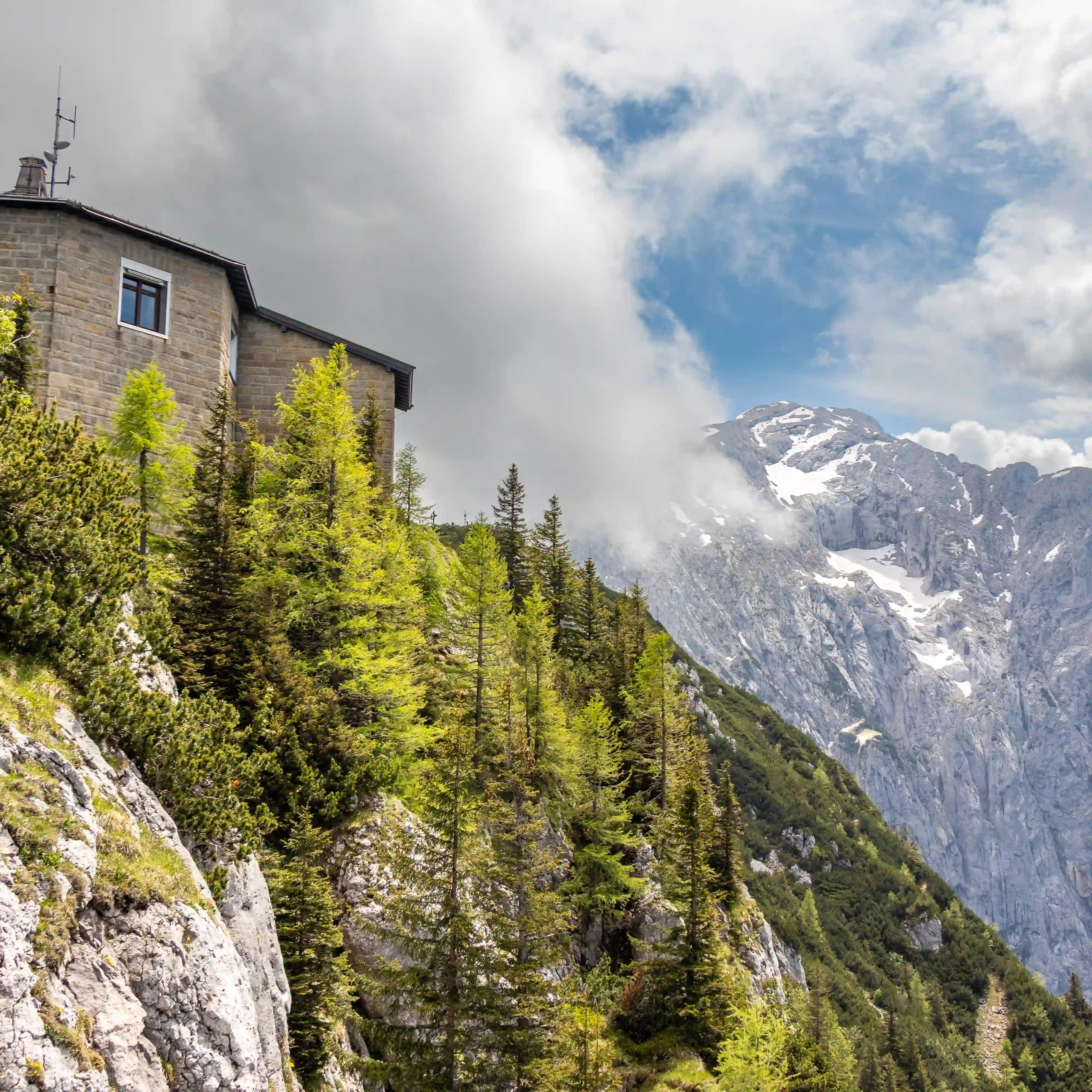 A stone building perched on a rocky slope with alpine trees and clouds drifting across nearby peaks.