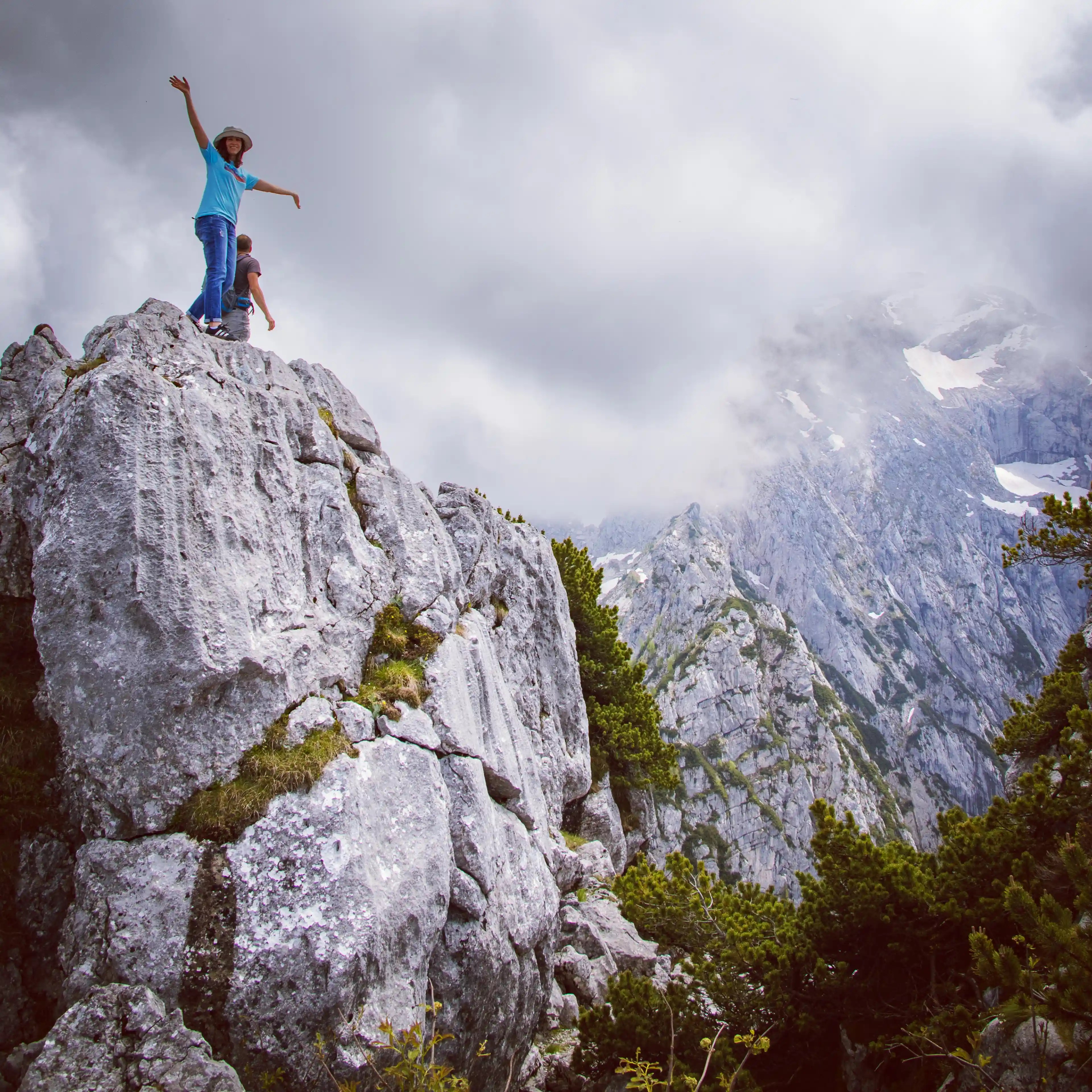 A person standing on a rocky outcrop with arms raised, surrounded by steep cliffs and alpine peaks.