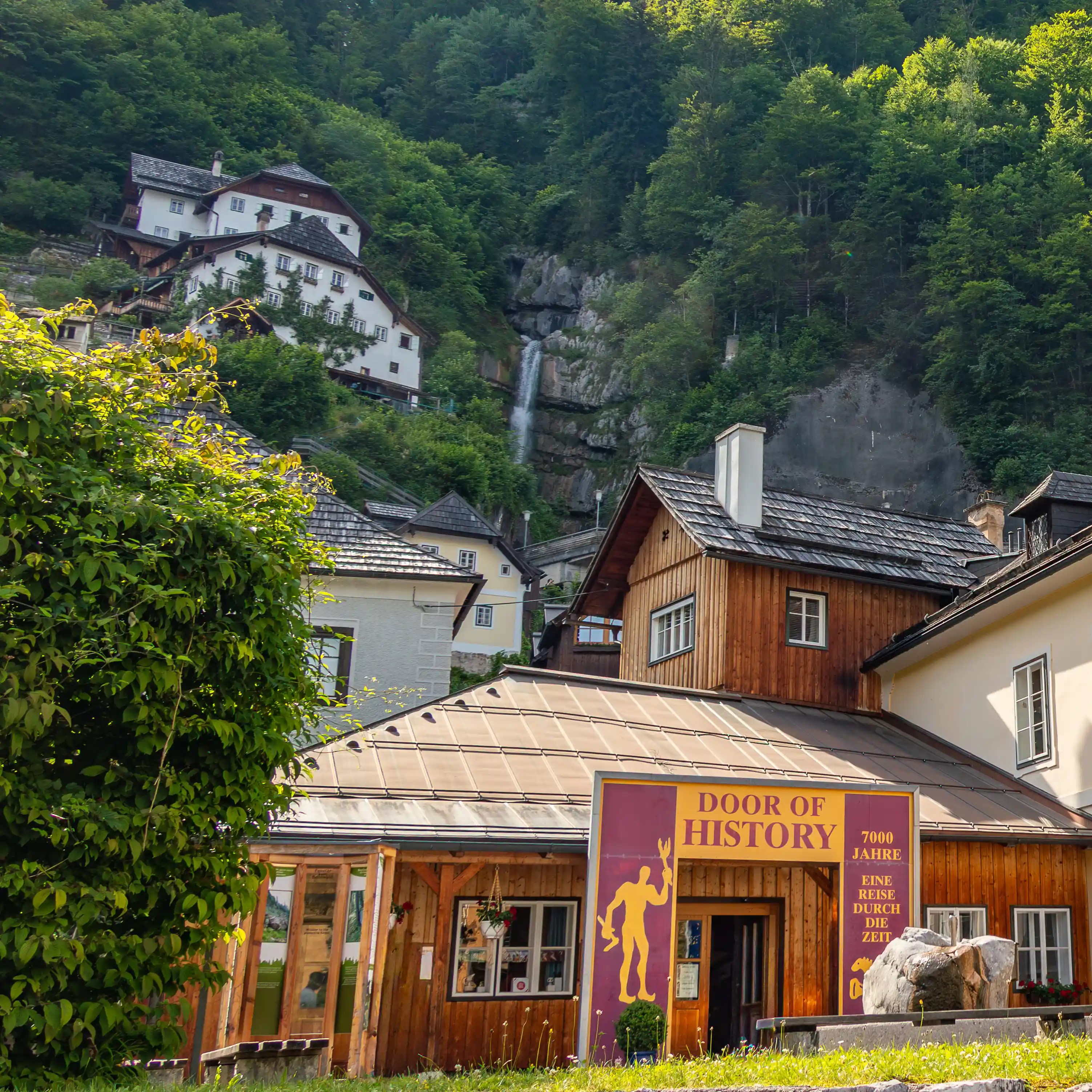 The Door of History museum building in Hallstatt with a waterfall cascading down the forested hillside behind it.