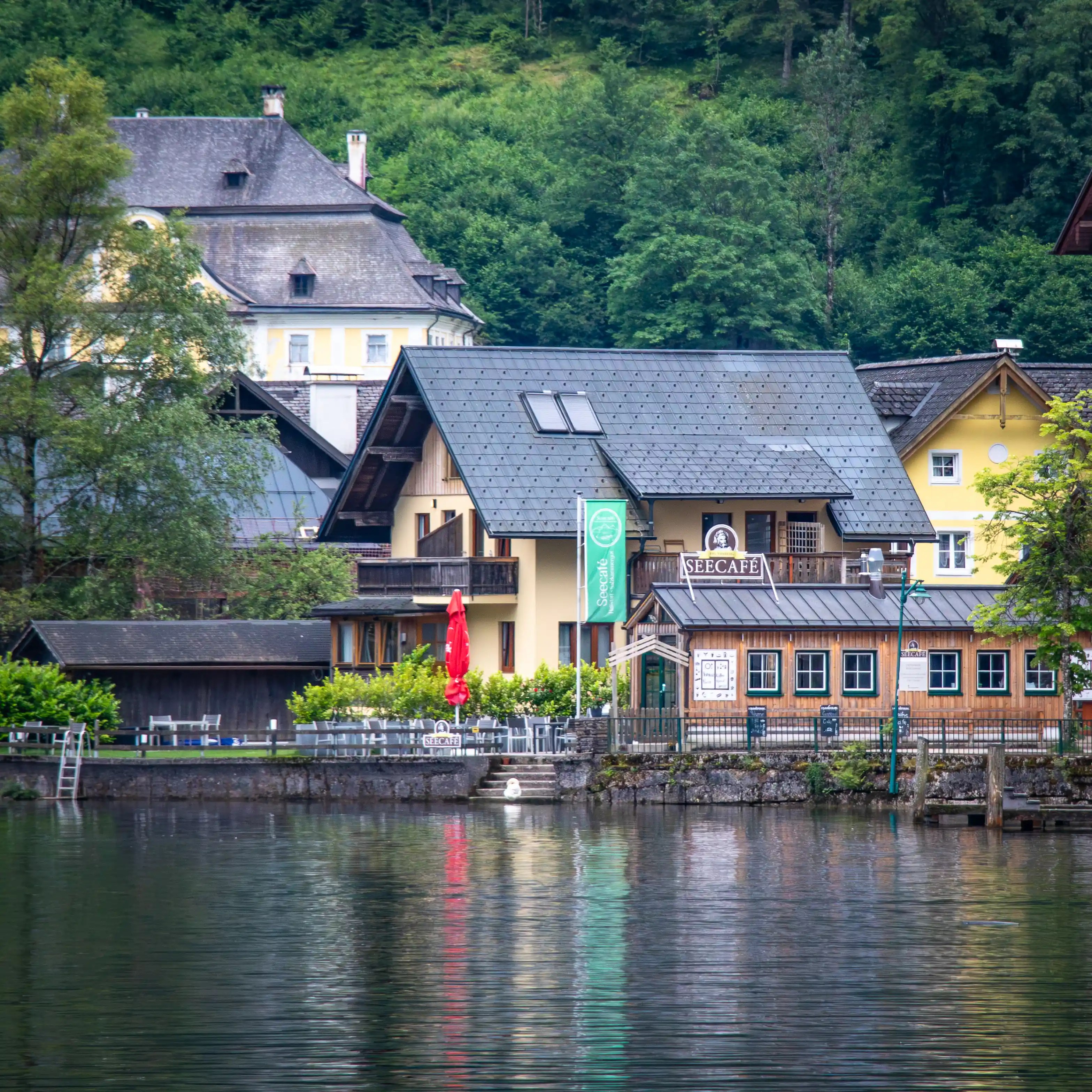 Lakeside guesthouse and cafe building in Hallstatt with reflected in the calm water of Lake Hallstatt, backed by a forested hillside.