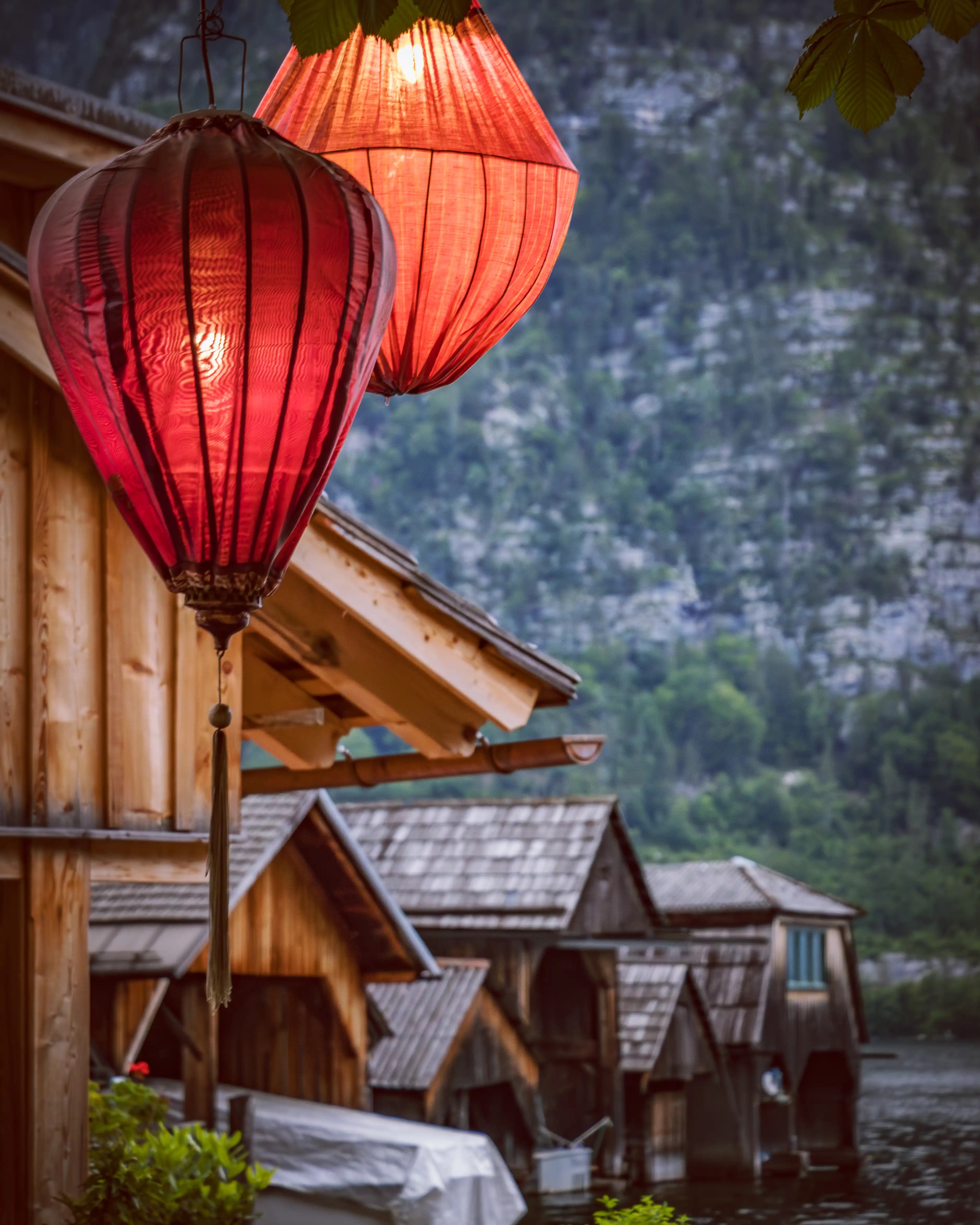Two glowing red and orange lanterns hang from a wooden lakeside tree in Hallstatt, with traditional boathouses and a forested mountain rising across the water in the background.