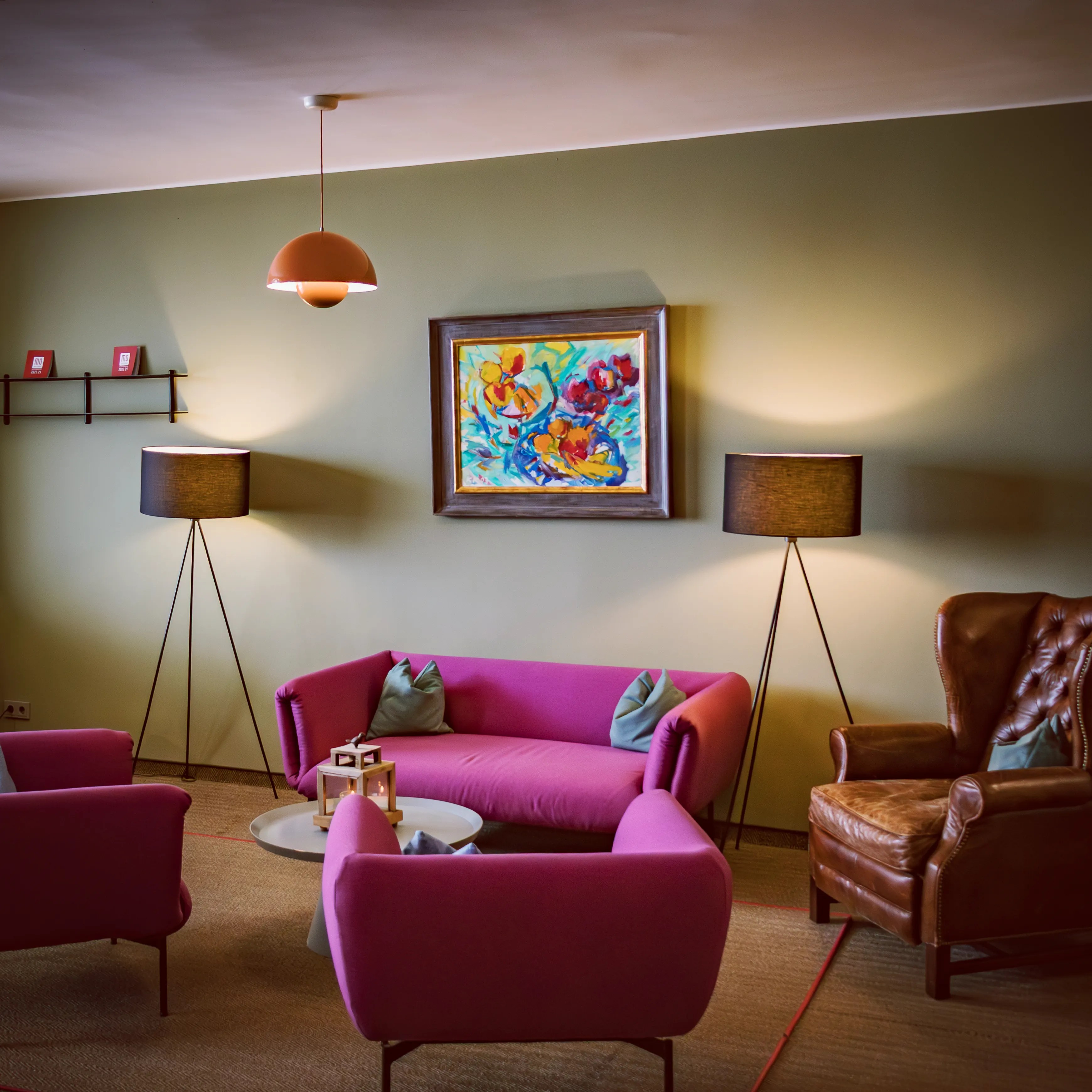 A cozy hotel lounge with magenta sofas, warm floor lamps, a leather armchair, and colorful artwork on a soft green wall.