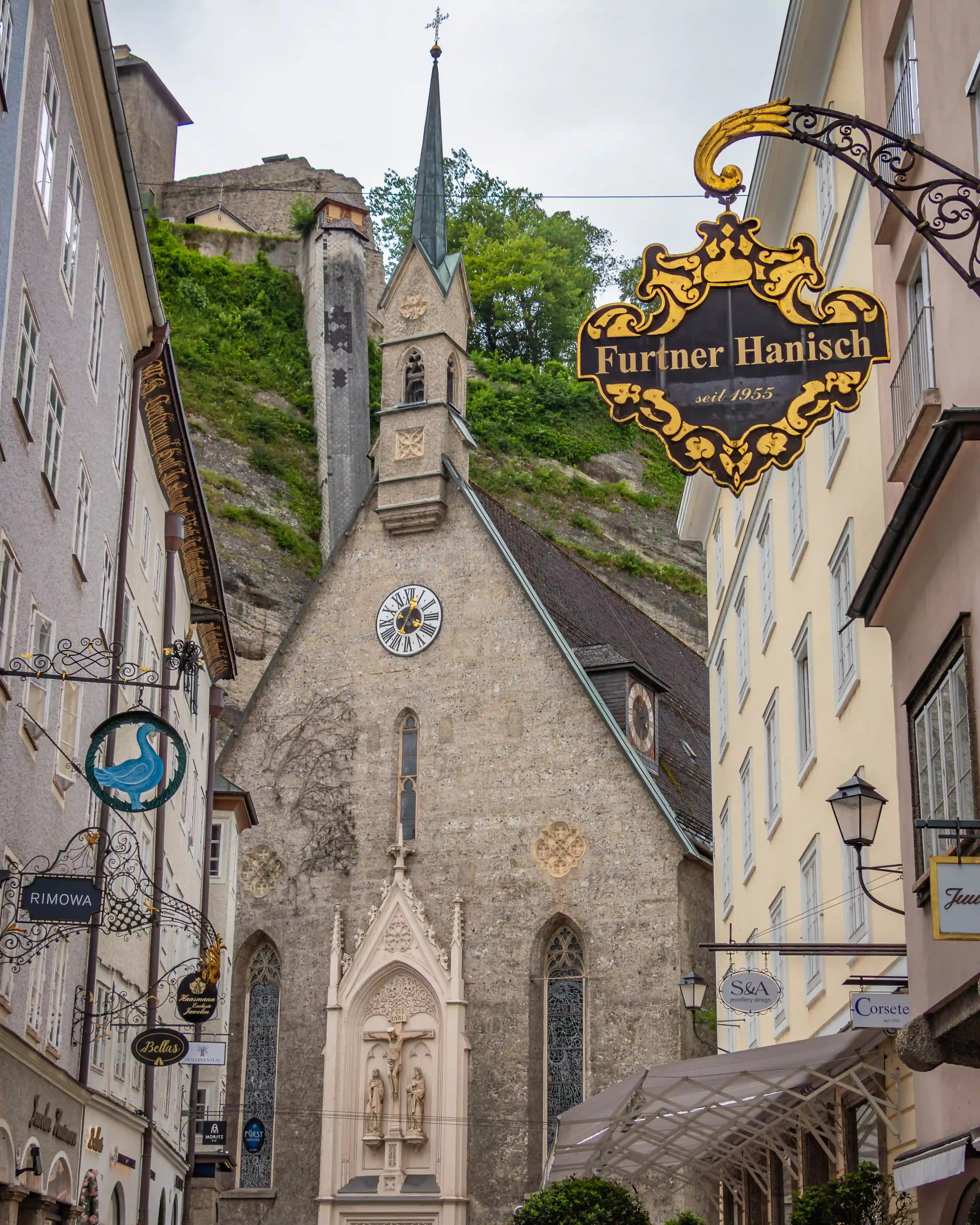 Kirche St. Blasius on Getreidegasse framed by historic buildings and shop signs in Salzburg Old Town.