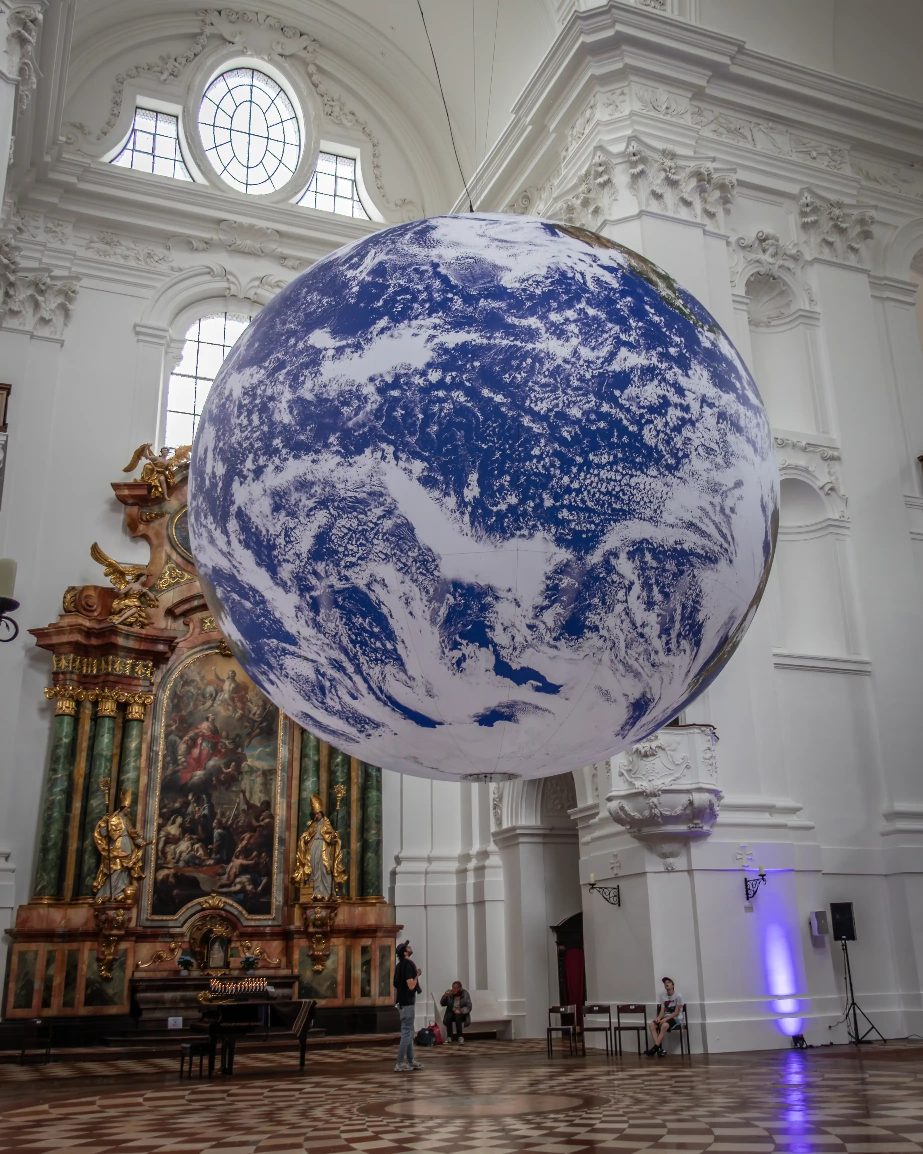 A large suspended globe depicting Earth hangs inside the bright white Baroque interior of the Salzburg Collegiate Church, with ornate stucco walls and an altar in the background.
