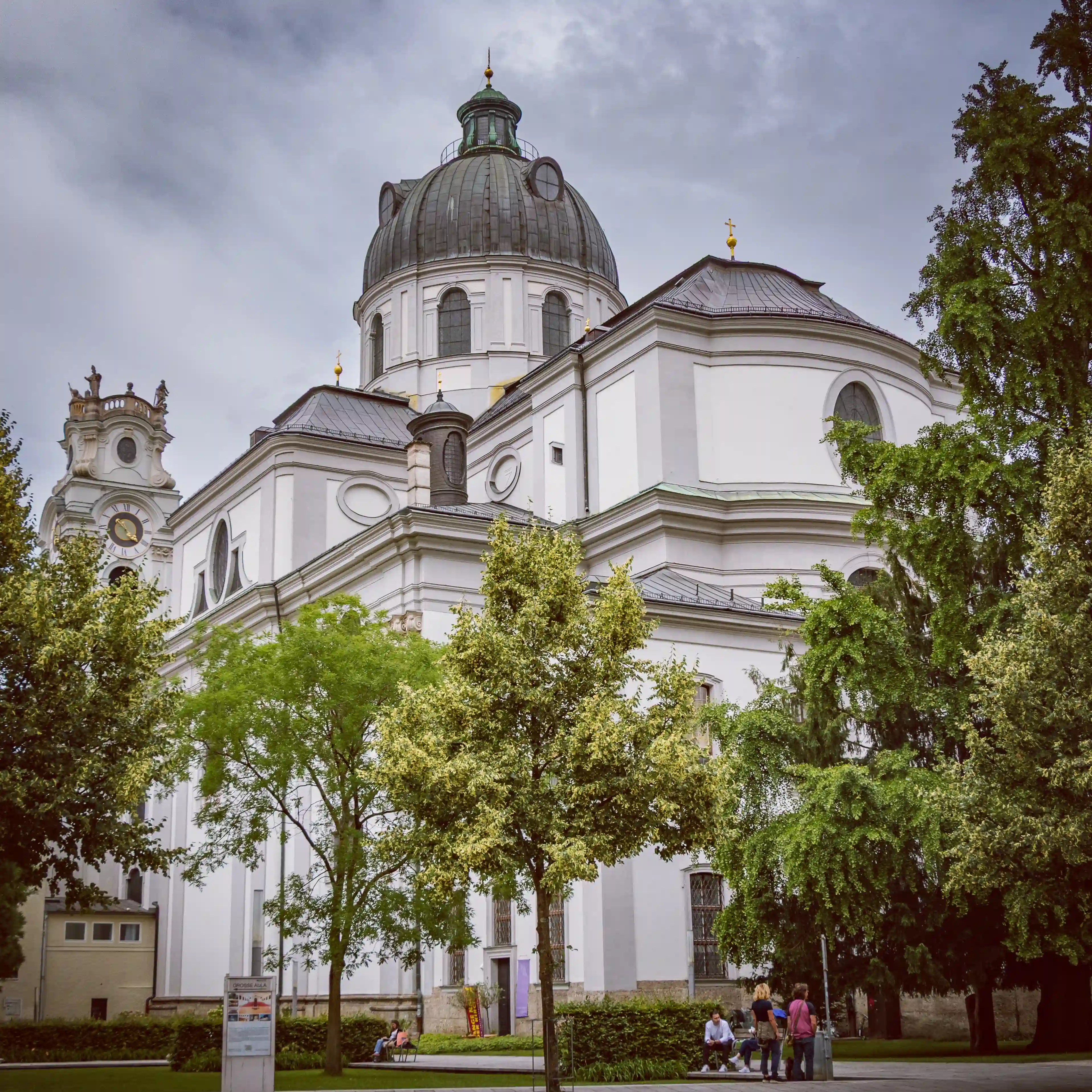 The white Baroque exterior and central dome of the Salzburg Collegiate Church rise above trees and pathways under a cloudy sky.