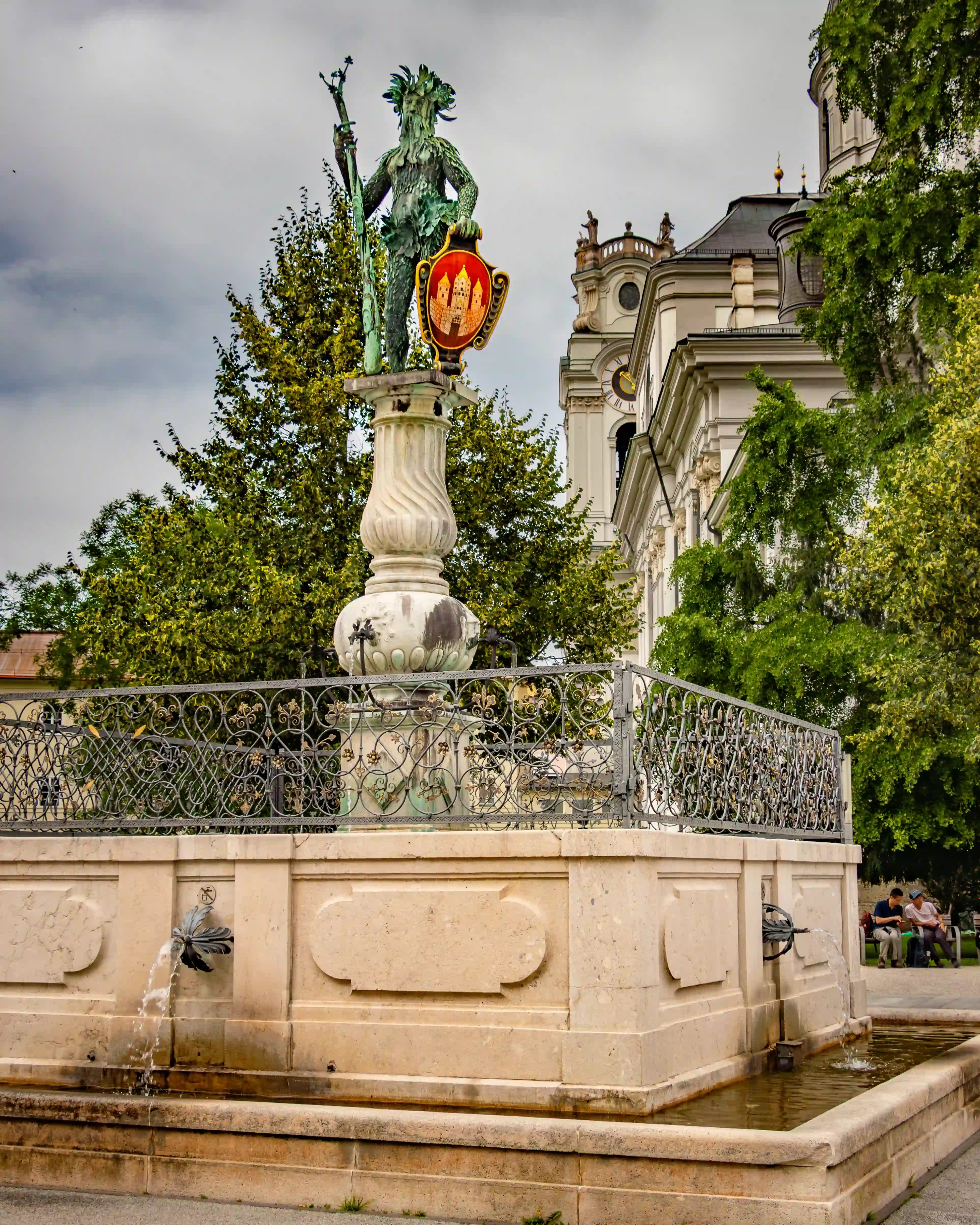 The Wilder-Mann Brunnen fountain features a bronze wild man statue holding a staff and the Salzburg coat of arms, set in front of the Salzburg Collegiate Church.