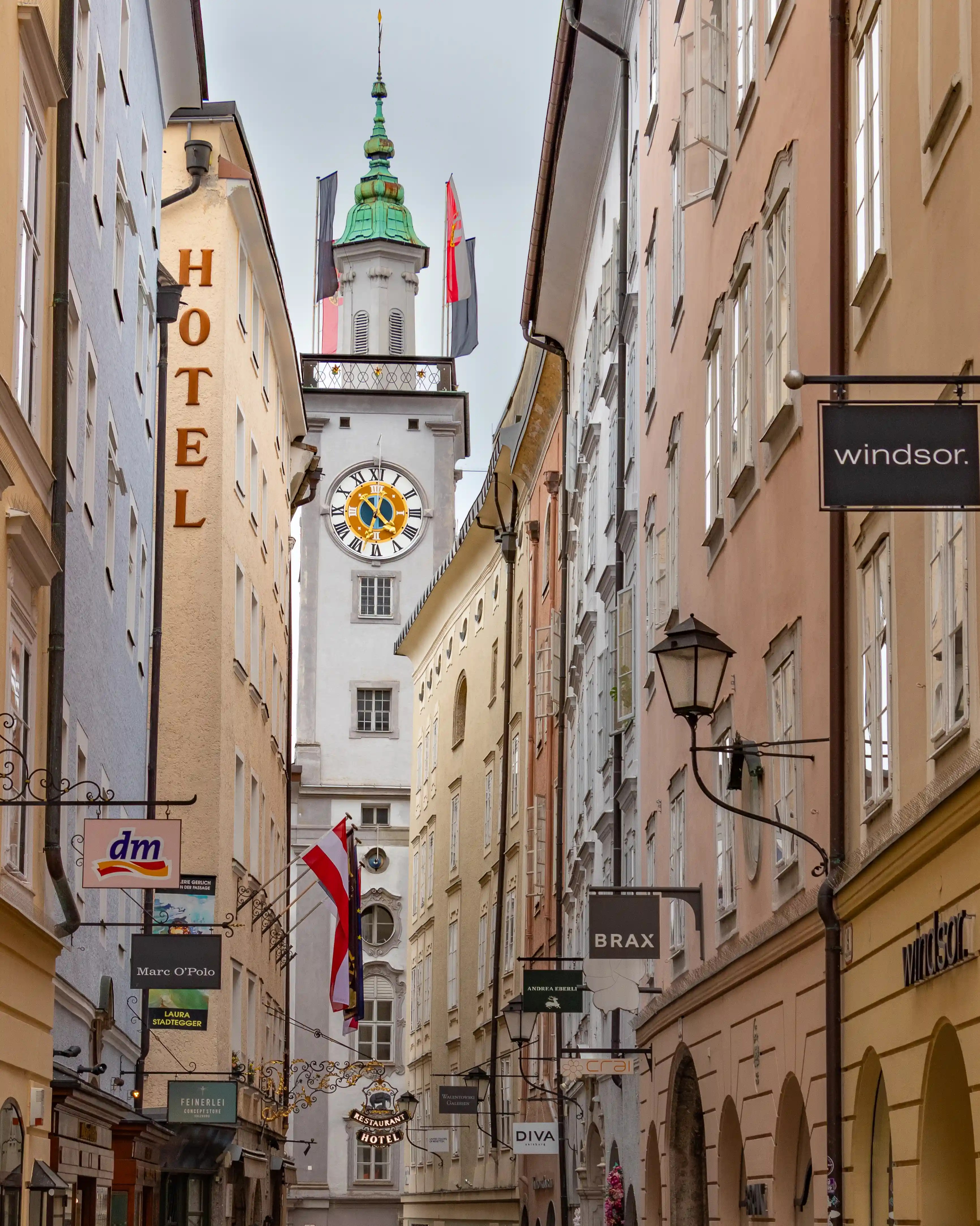 Narrow street in Salzburg’s Old Town lined with tall pastel buildings and hanging shop signs, leading toward the white Old Town Hall clock tower with a green spire and clock face.