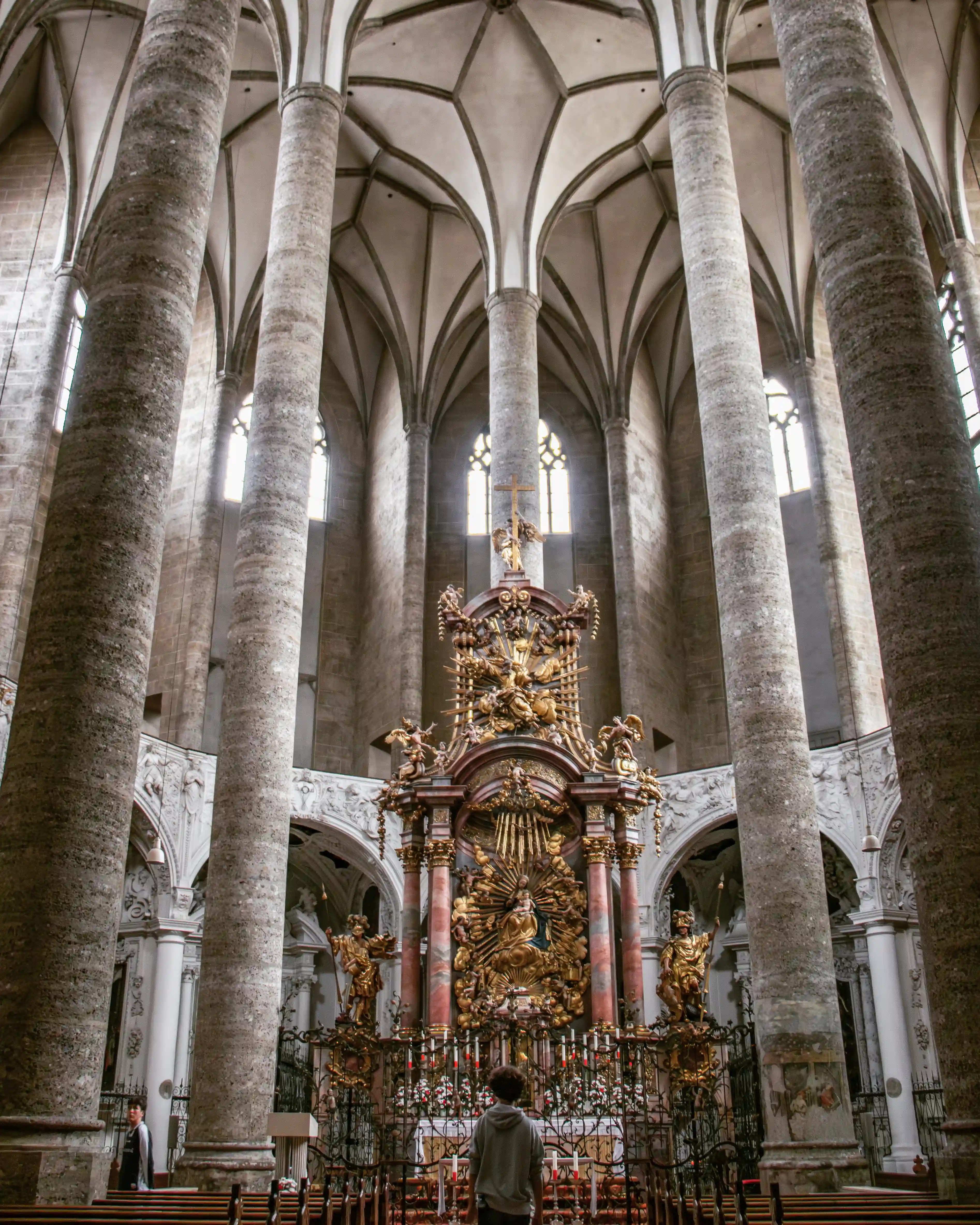 A tall Gothic nave with stone columns rises toward a vaulted ceiling, framing an ornate gilded altar at the center of Salzburg’s Franciscan Church.