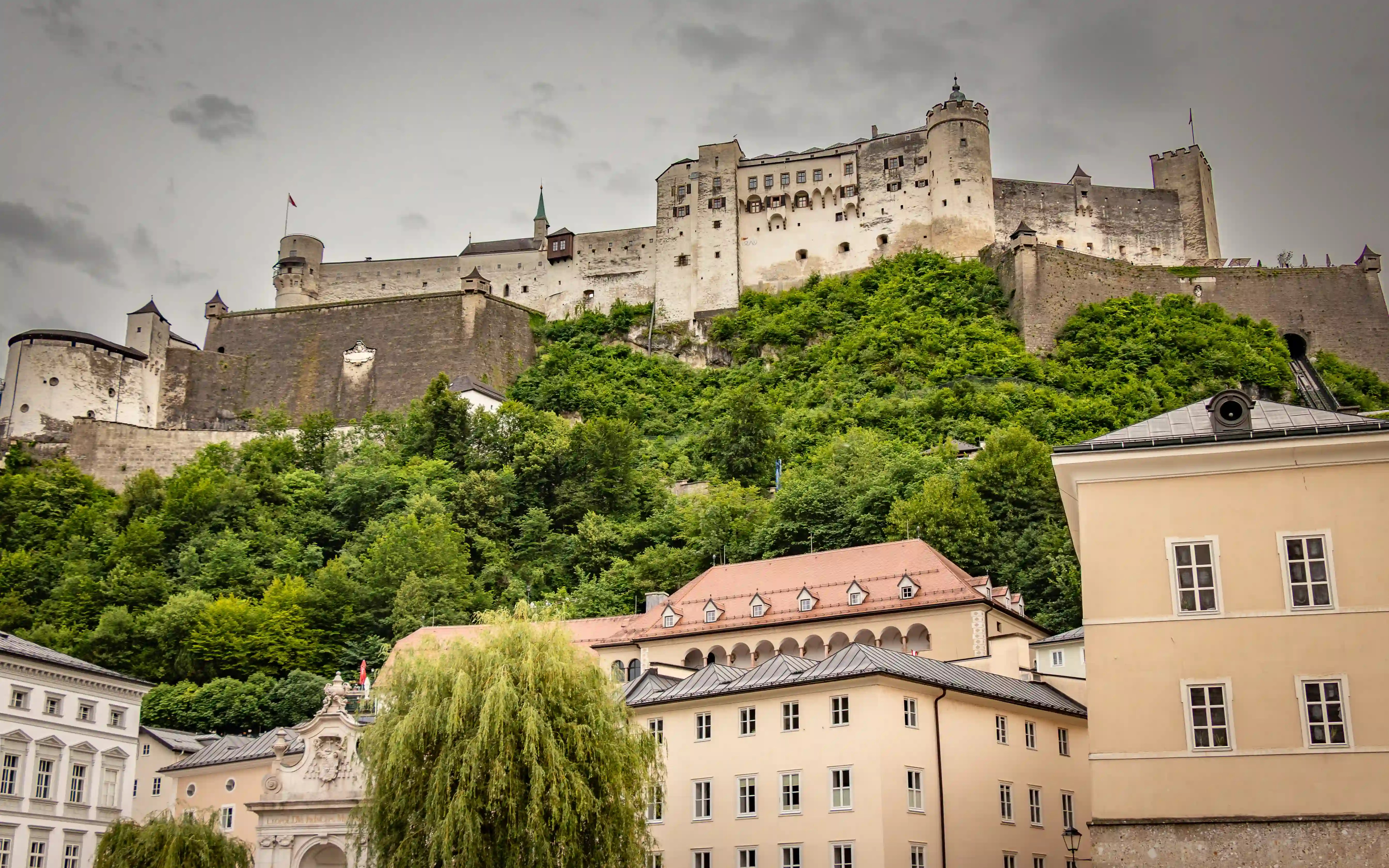 Massive stone fortress walls and towers of Hohensalzburg Fortress rising above green hills and pale historic buildings in Salzburg.