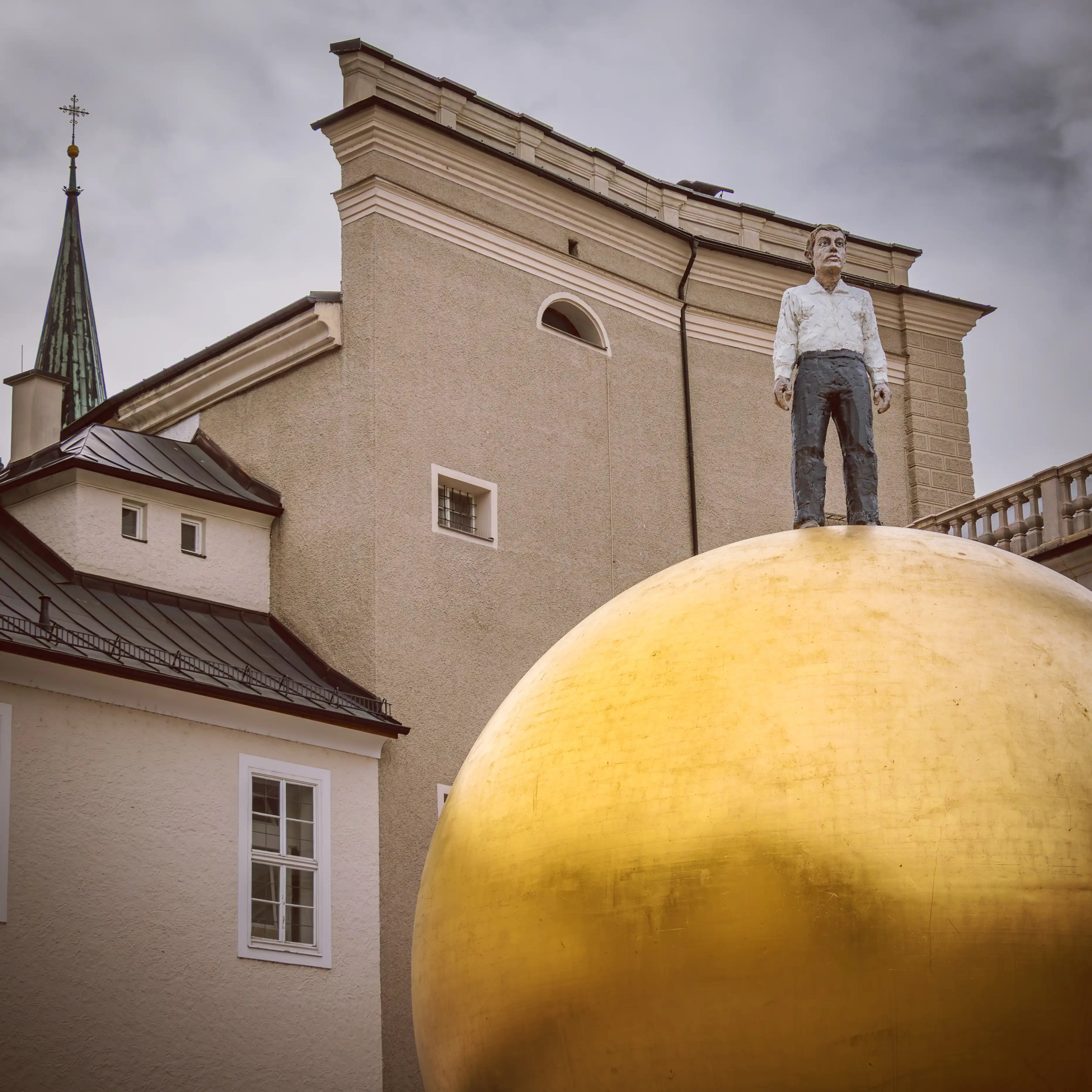 A statue of a man standing atop a large golden sphere in Kapitelplatz, with a historic building behind it.