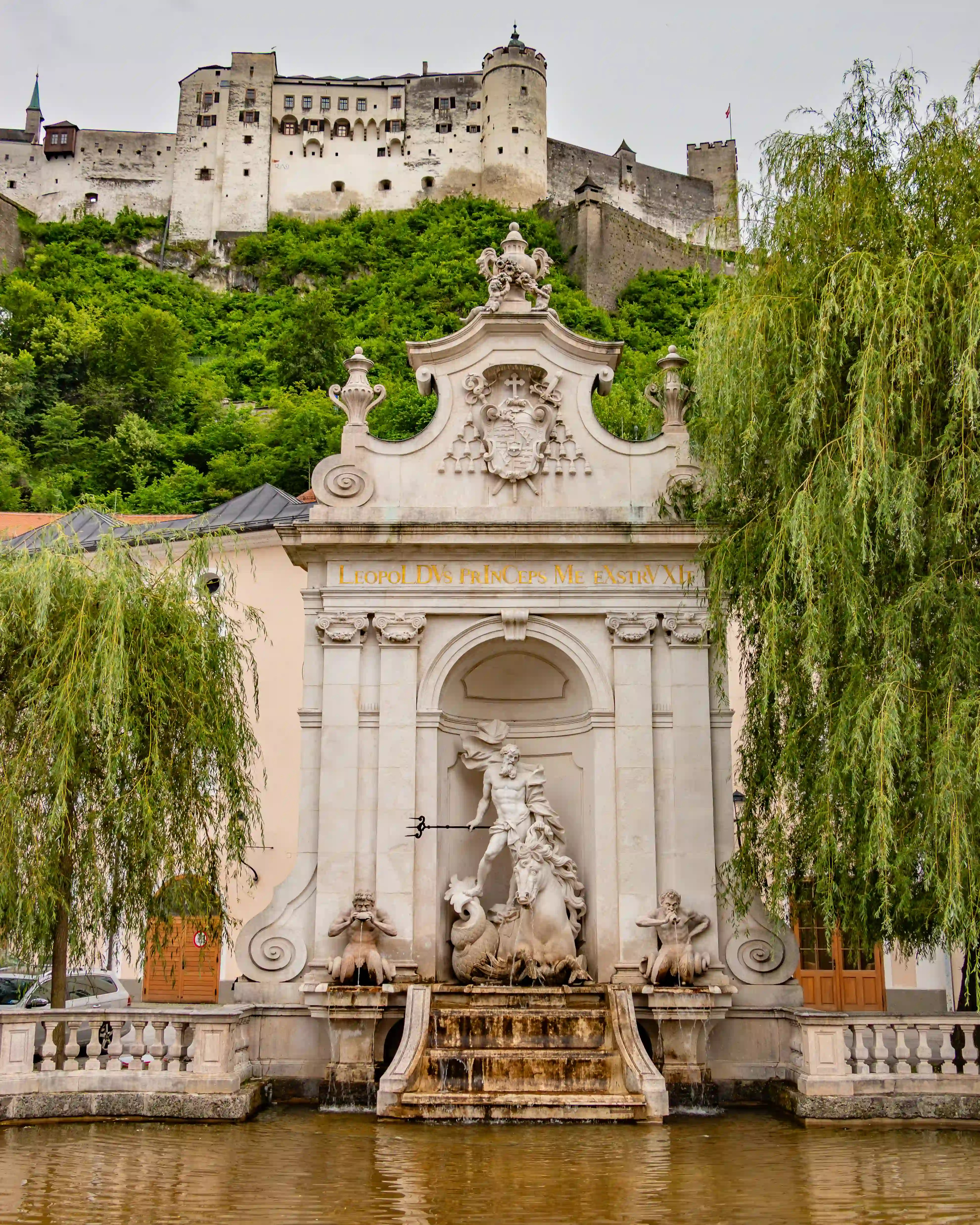 Large Baroque horse trough fountain with a sculpted Neptune figure standing in an arched stone niche, water flowing into a shallow basin, with Hohensalzburg Fortress rising on the green hillside behind.