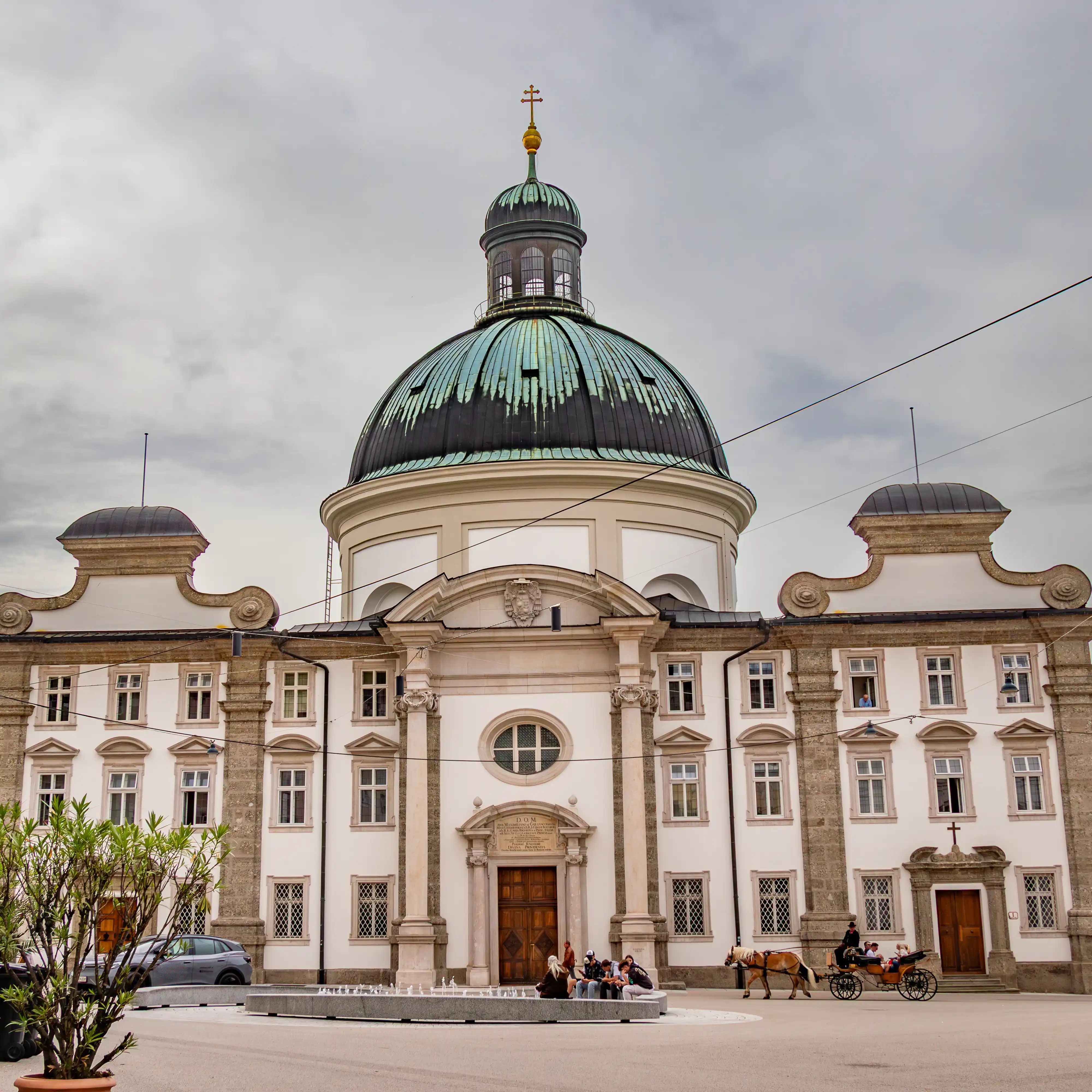 Baroque church with a large green copper dome and white façade with people sitting near a low fountain and a horse-drawn carriage passing in front.