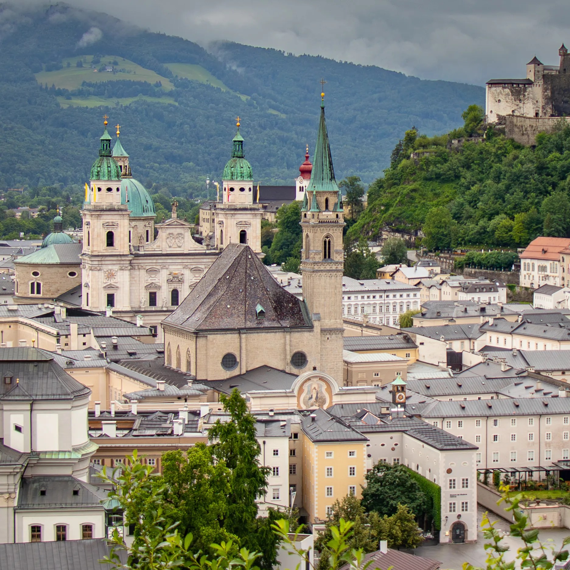 A cityscape view of Salzburg shows the towers of Salzburg Cathedral and the Franciscan Church rising above pastel buildings with forested hills in the background.