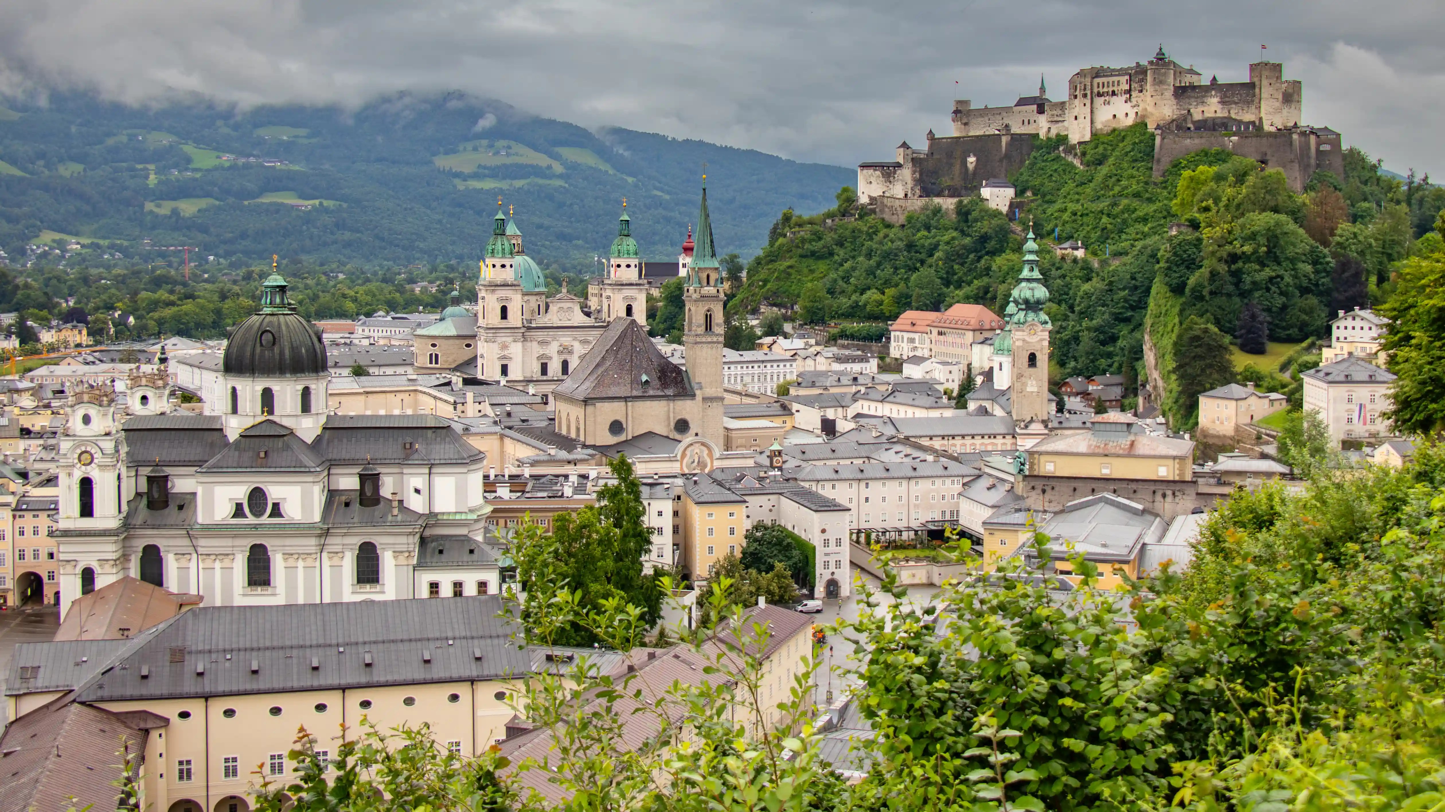 A panoramic view of Salzburg’s Old Town with multiple church towers, green hills, and Hohensalzburg Fortress on the ridge in the background.