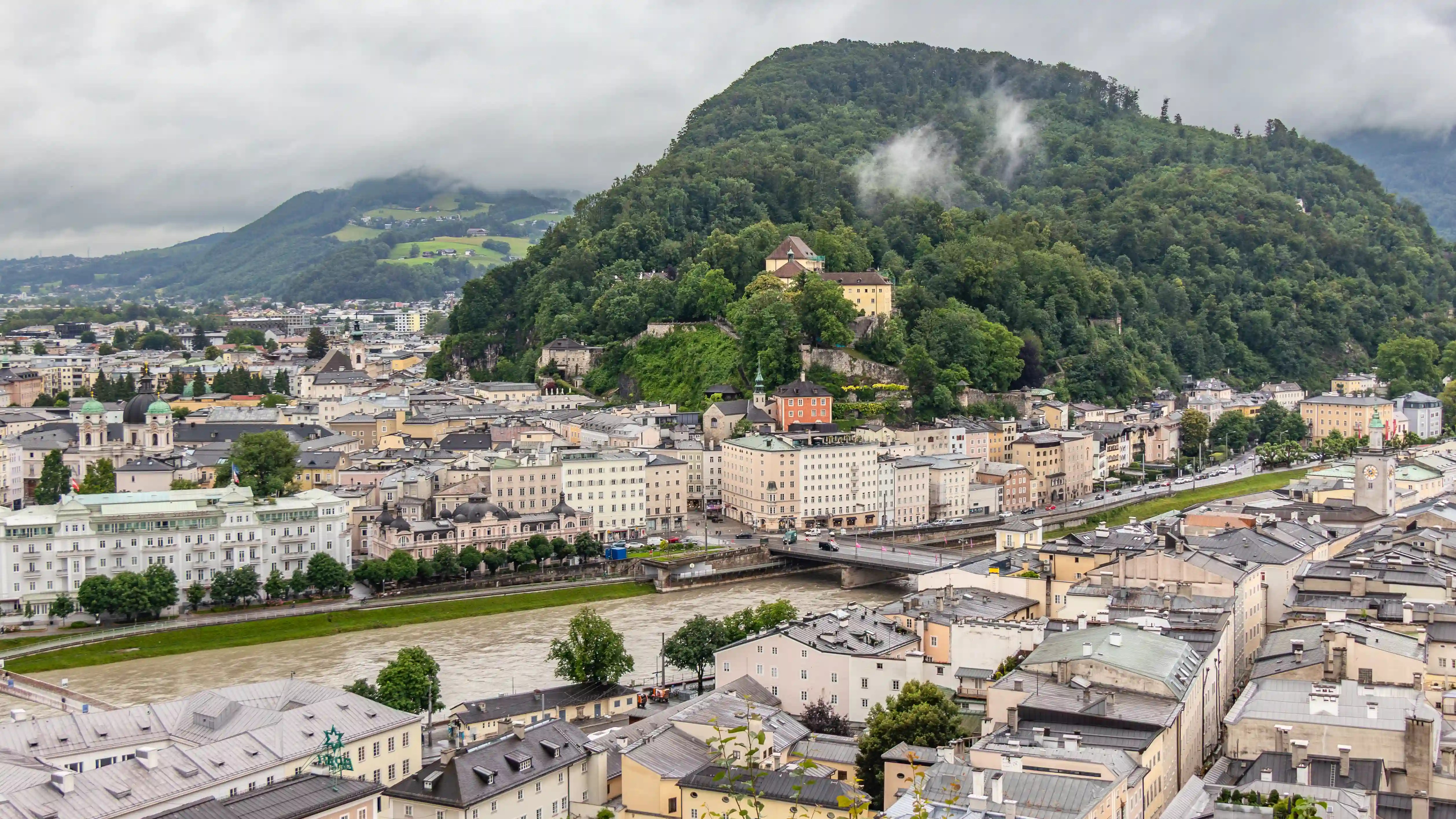 A city view along the Salzach River with bridges, historic buildings, and a tree-covered hill rising behind the Old Town.
