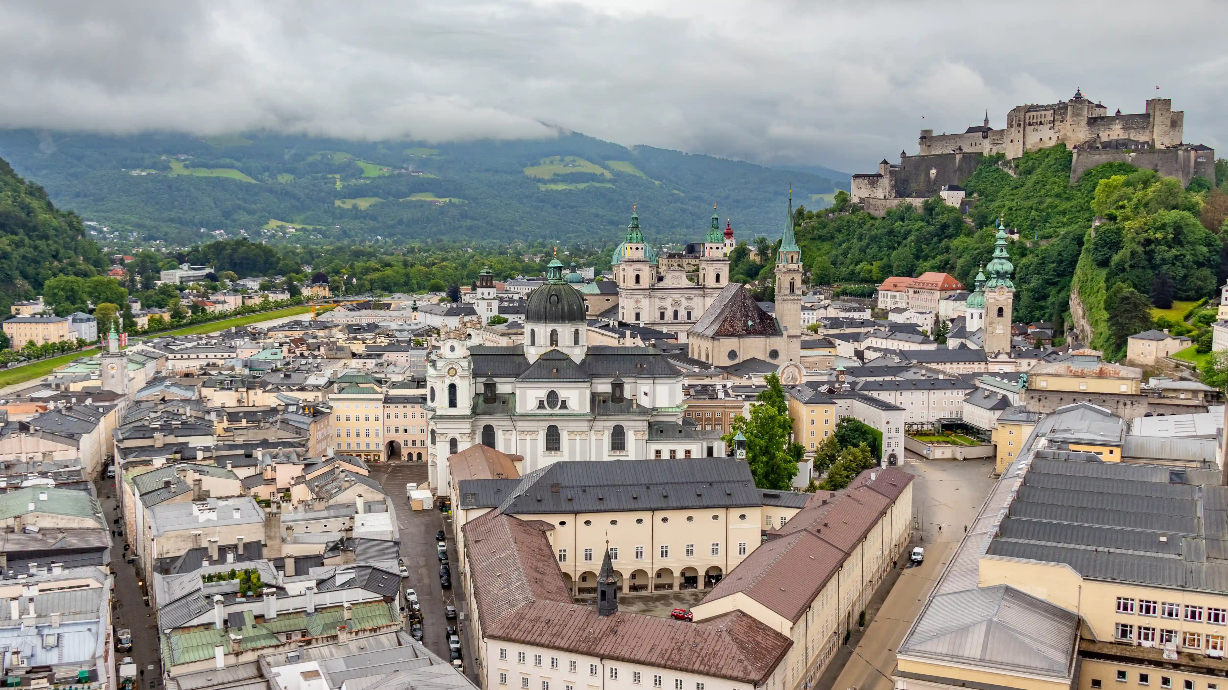 A wide view over Salzburg’s Old Town with church domes and towers below and Hohensalzburg Fortress rising on the forested hill in the distance under a cloudy sky.