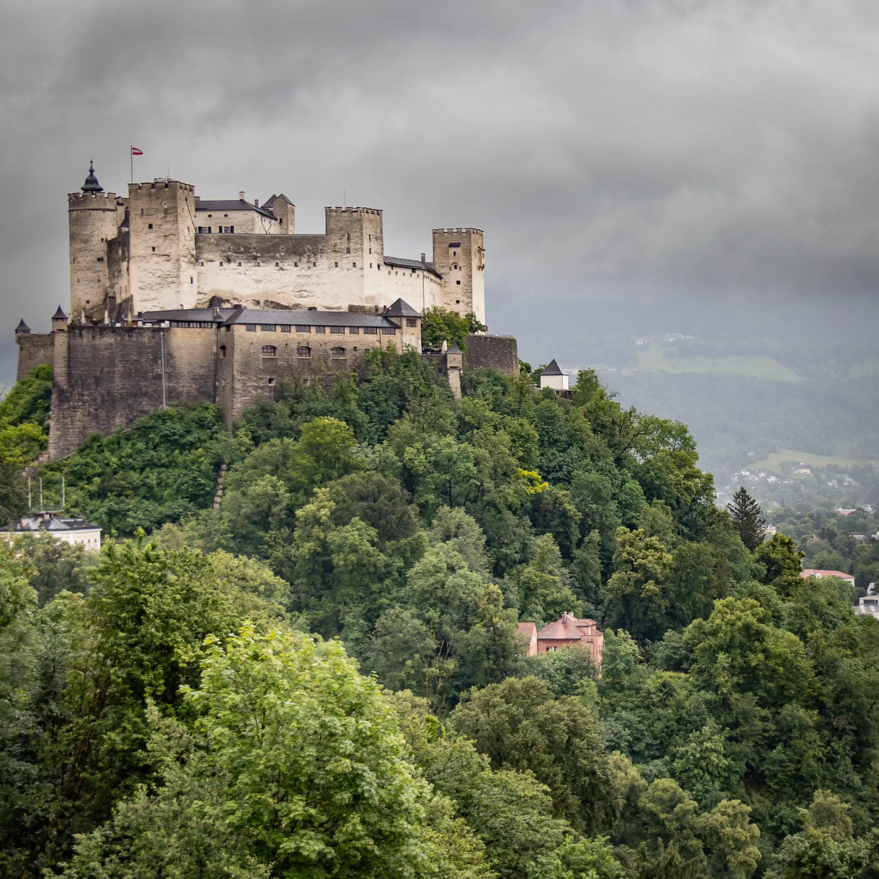 Hohensalzburg Fortress rising above dense forest on a steep hill under an overcast sky.