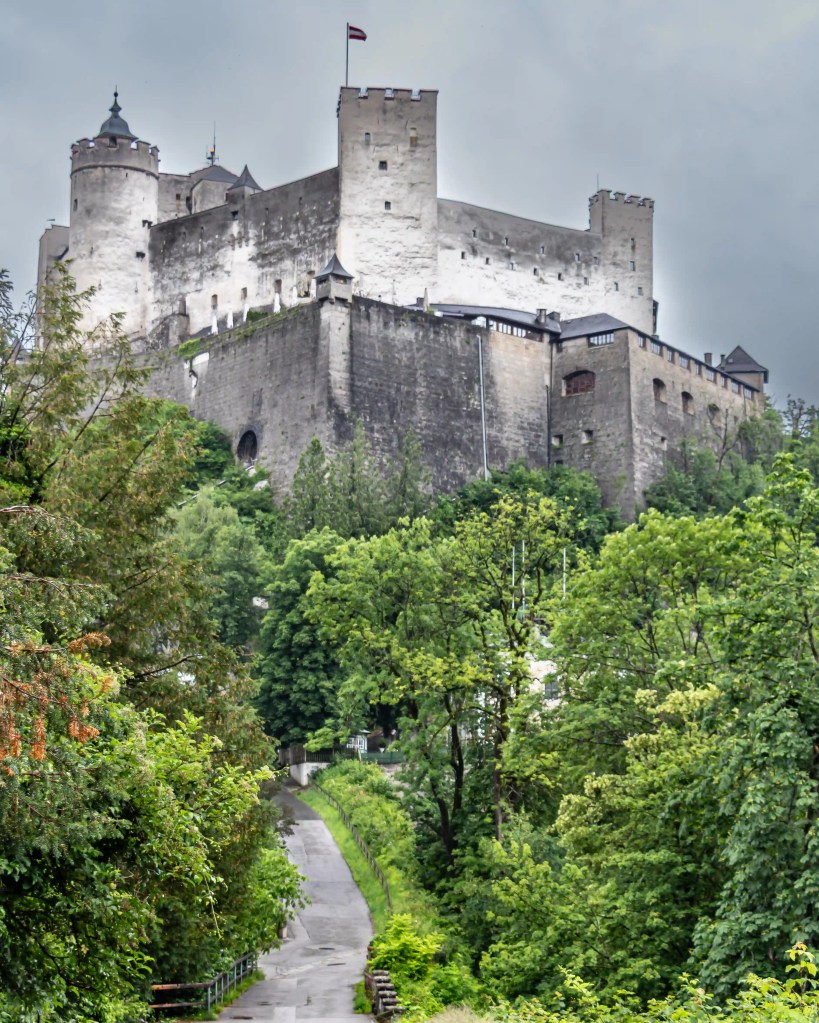 A massive white stone fortress rises above a steep, tree-lined hillside, with a narrow paved path winding upward through dense green foliage.