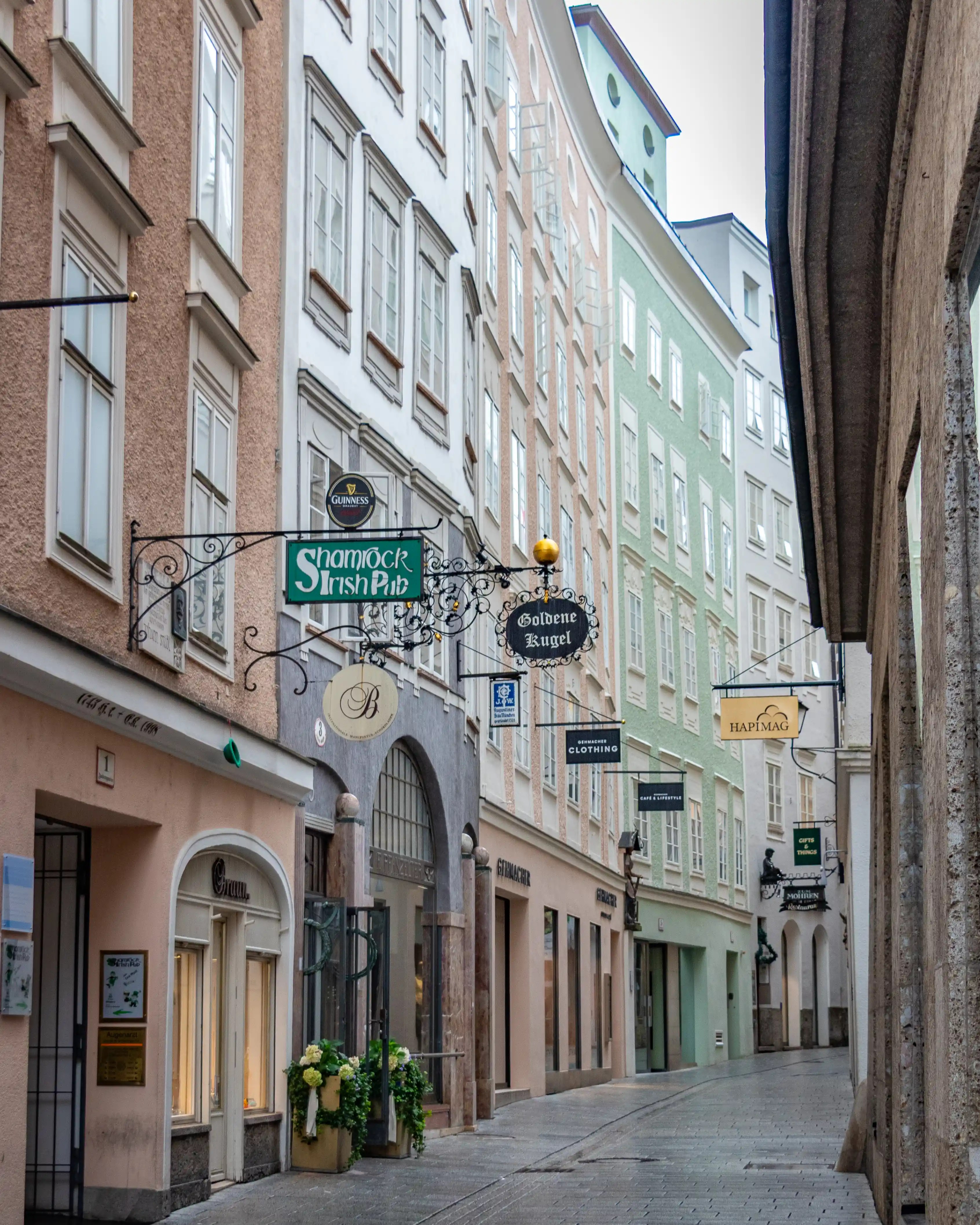 A narrow pedestrian street in Salzburg with pastel buildings, ornate wrought-iron shop signs, and a gently curving stone pavement.