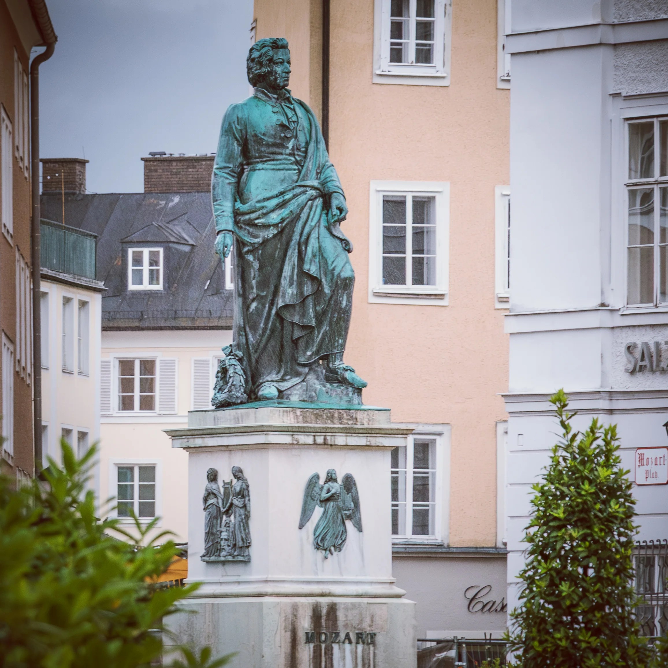 A bronze statue of Wolfgang Amadeus Mozart standing on a stone pedestal in a Salzburg square, surrounded by historic buildings.