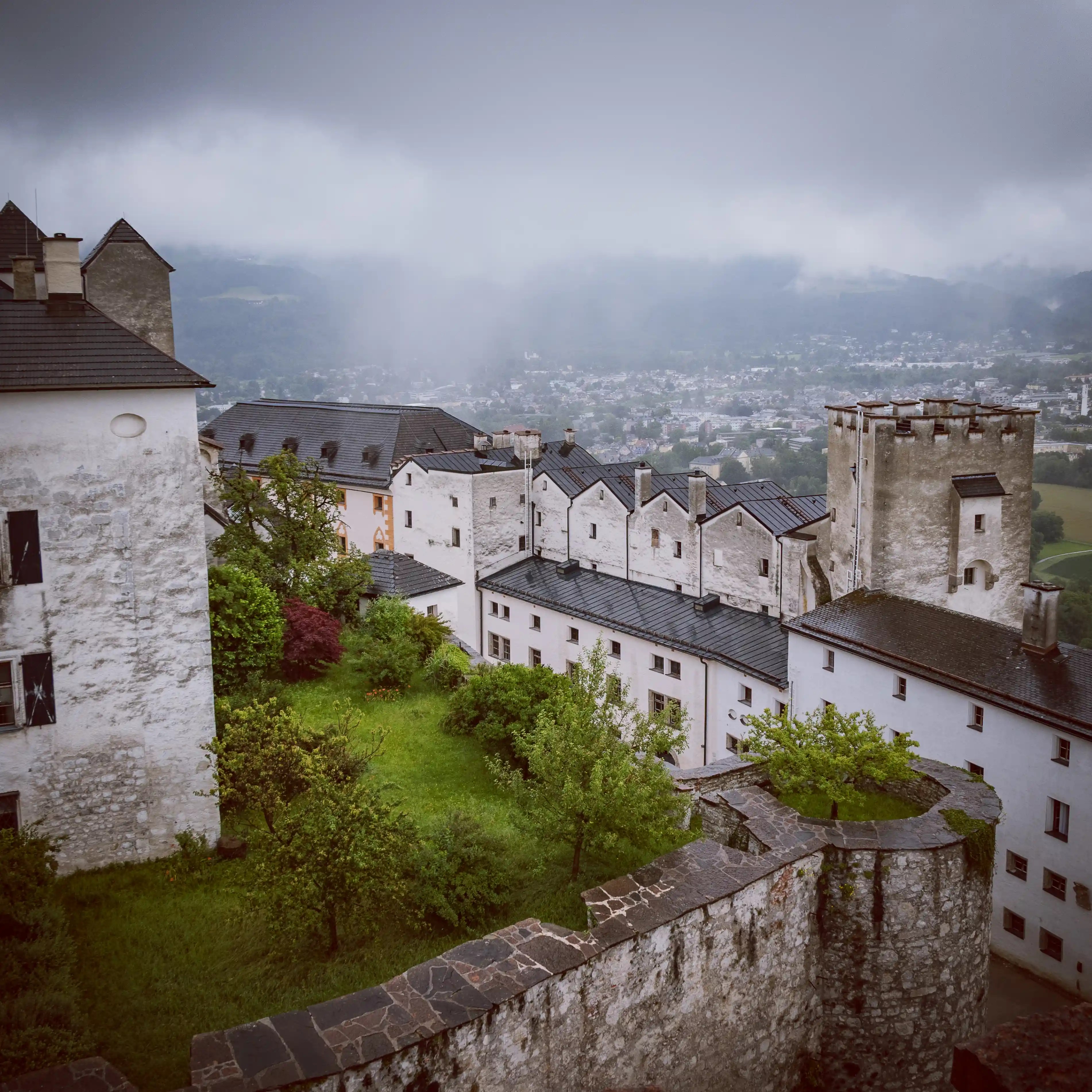 White fortress buildings with dark roofs surround a grassy courtyard, with Salzburg and misty hills visible in the distance.