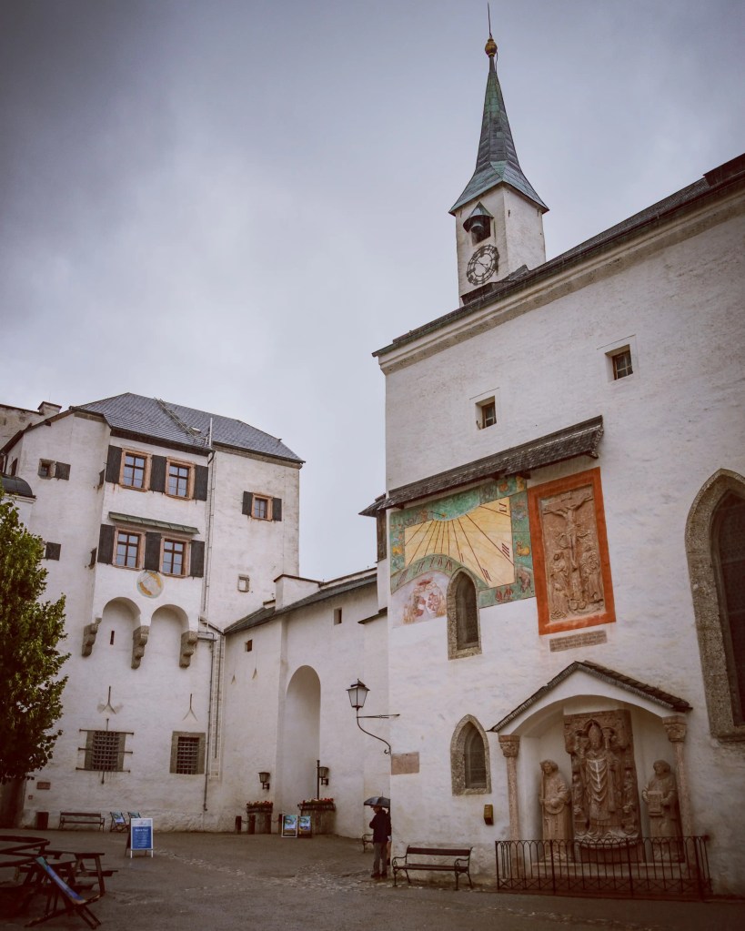 A small courtyard features white stone buildings, a clock tower, and a painted sundial on the wall.