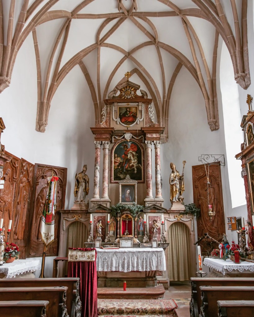 An ornate church altar stands beneath a vaulted ceiling, framed by statues, candles, and religious artwork.