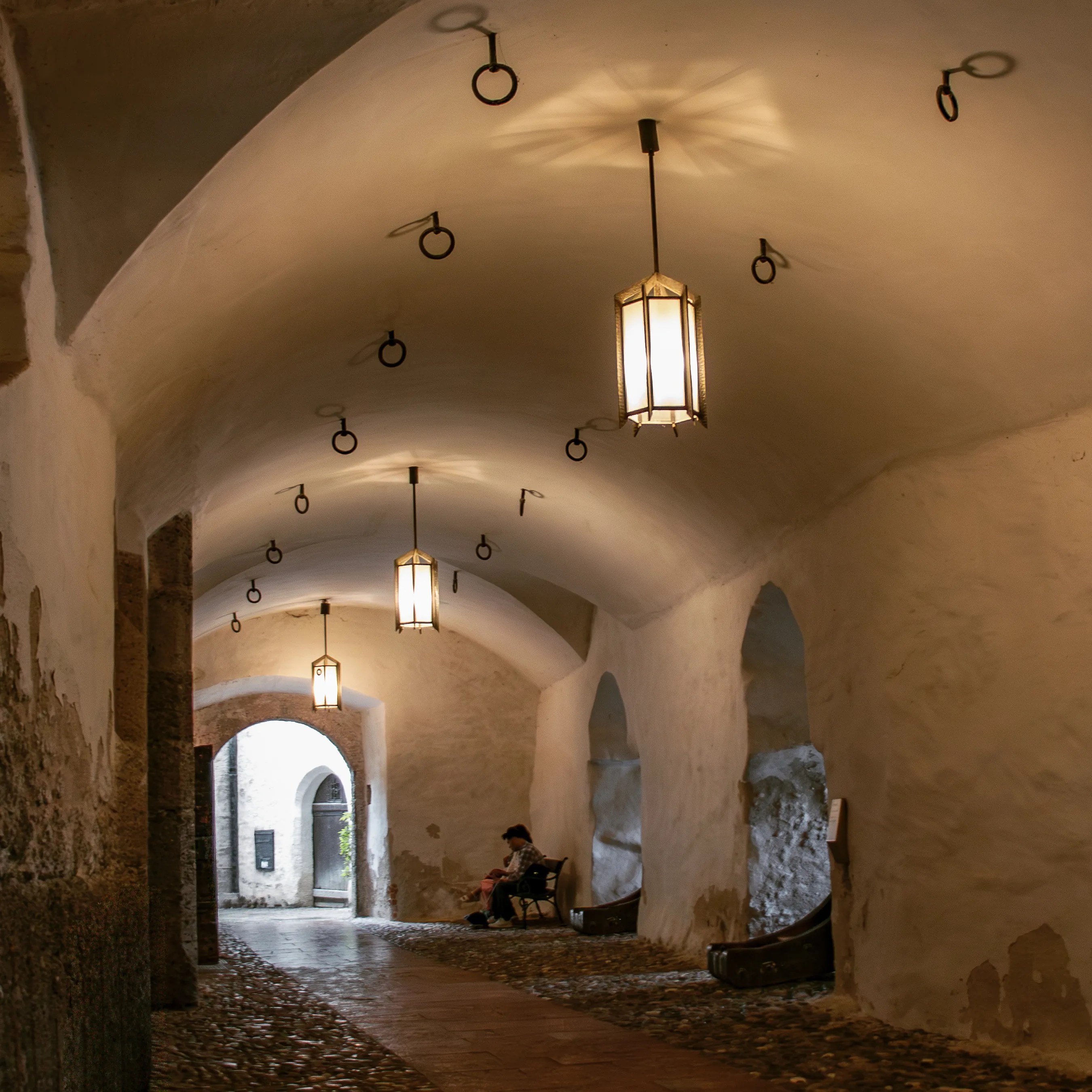 A vaulted stone passageway with hanging lanterns leads toward a bright arched exit, with benches along the walls.