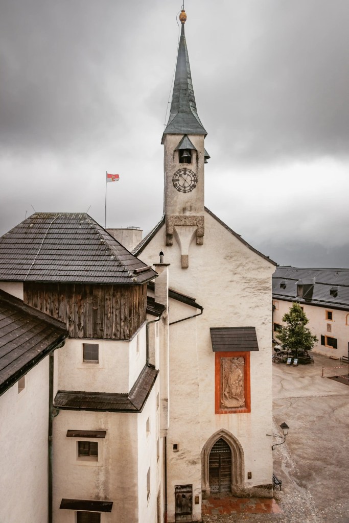 A white stone building with a narrow clock tower stands beside connected fortress structures under a gray sky.