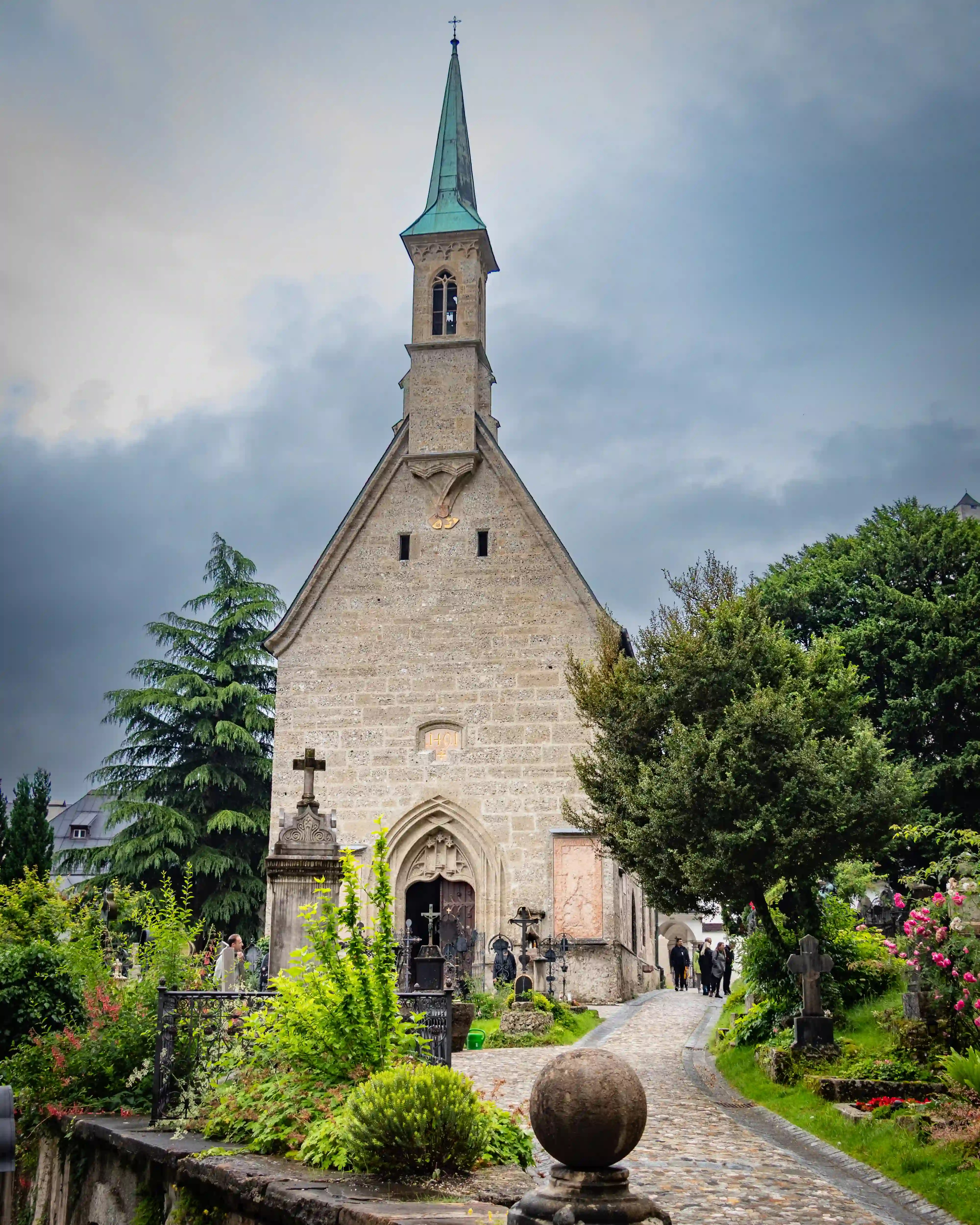 A narrow cobblestone path leads past gravestones toward a tall stone church building at St. Peter’s Cemetery in Salzburg.