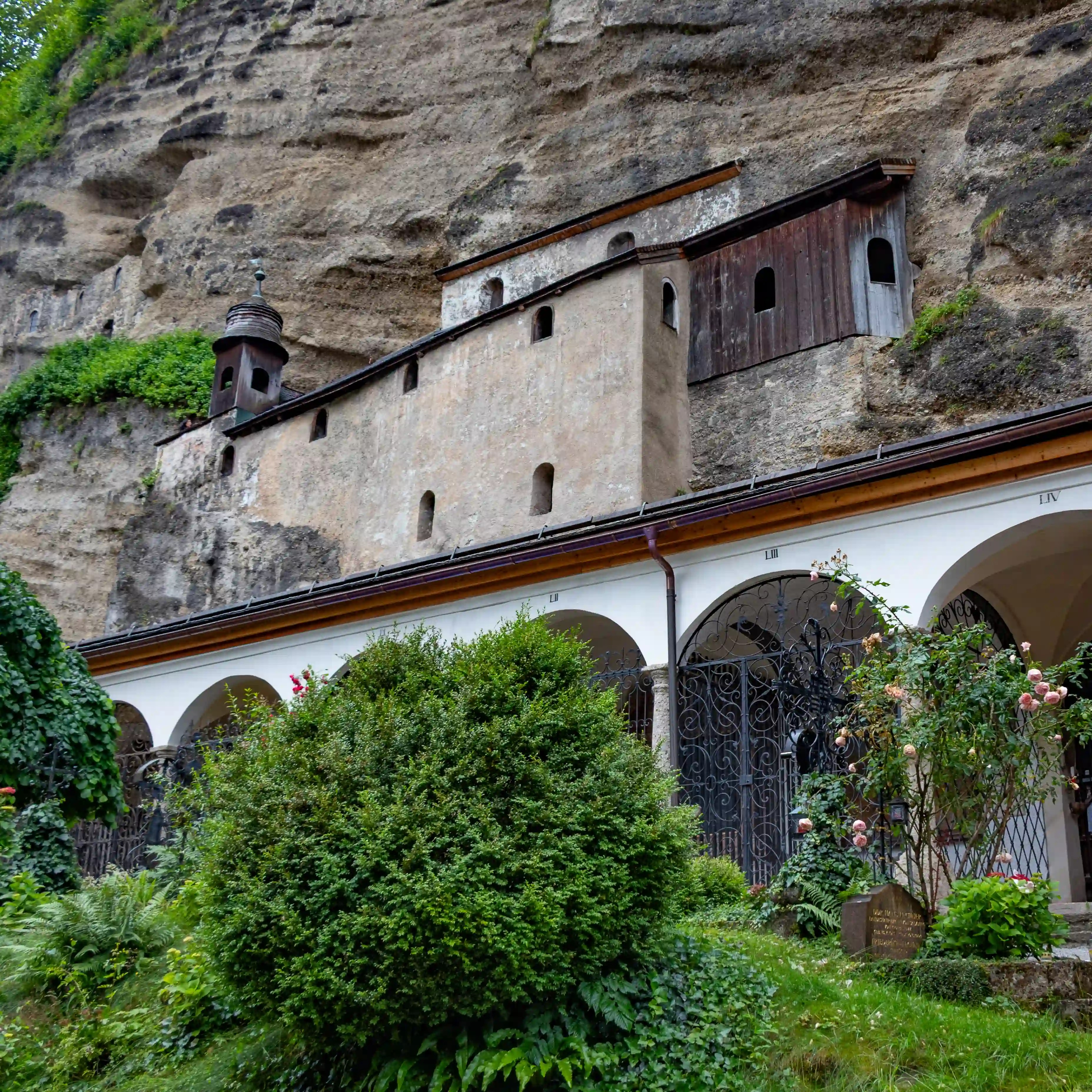 A long stone building with memorial plaques and small windows is built against the rock face at the St. Peter’s Catacombs in Salzburg.