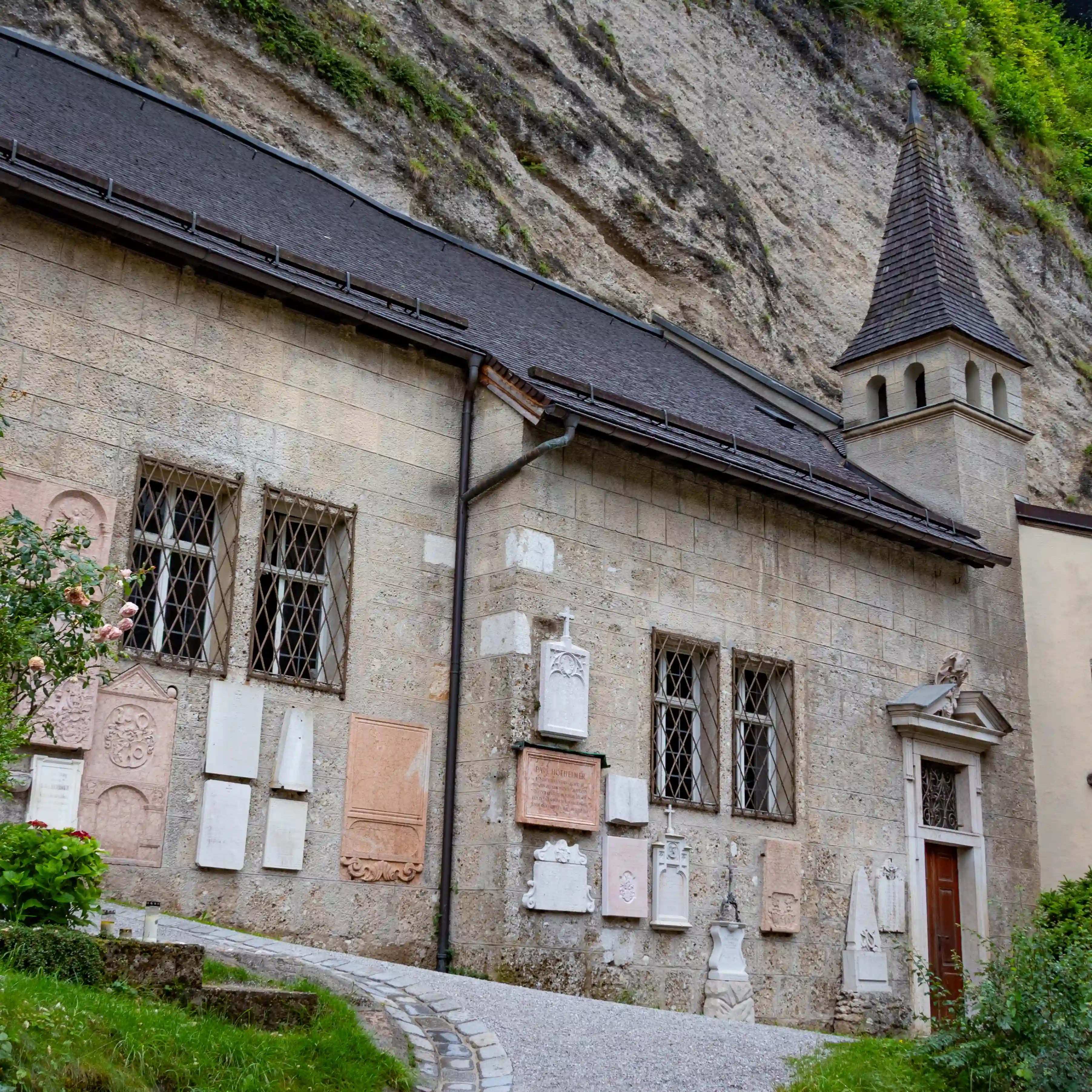 A stone church wall with small windows and a pointed steeple stands at the base of a rocky cliff near St. Peter’s Cemetery.