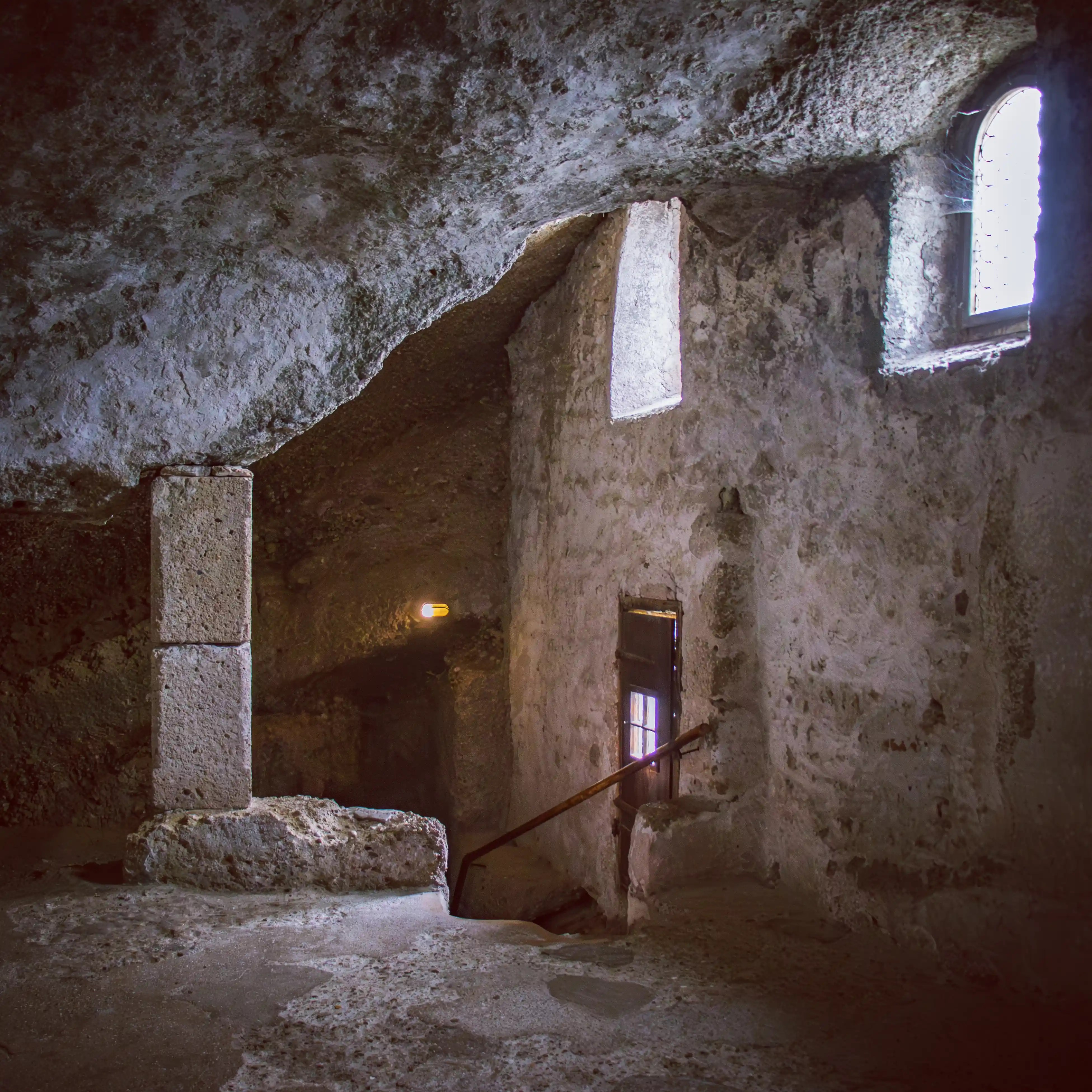 Stone-carved interior of the St. Peter’s Catacombs with a rough rock ceiling, a narrow staircase, and small windows letting in soft natural light.