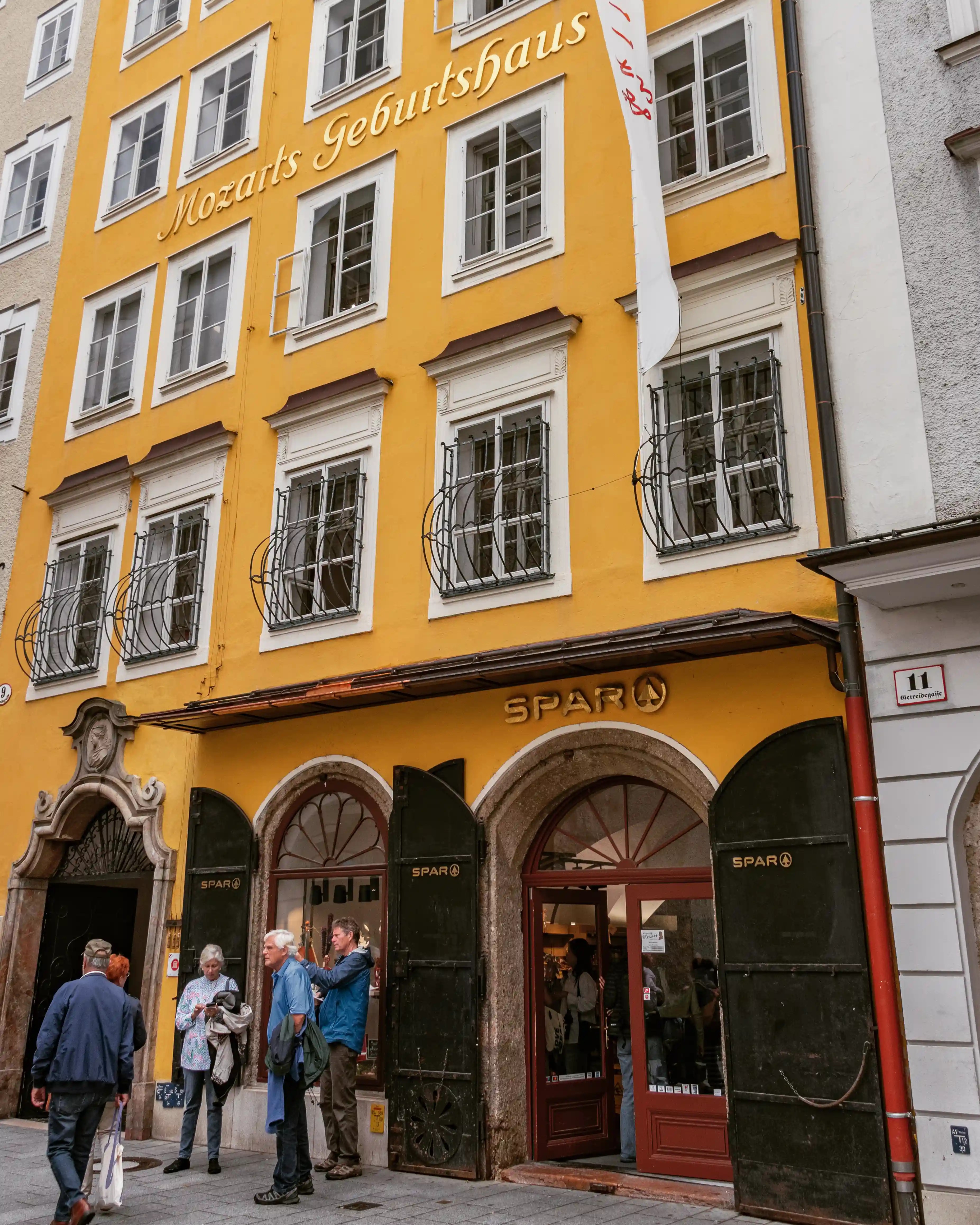 The bright yellow exterior of Mozarts Geburtshaus in Salzburg with white-trimmed windows, iron window grilles, and visitors gathered at the entrance.