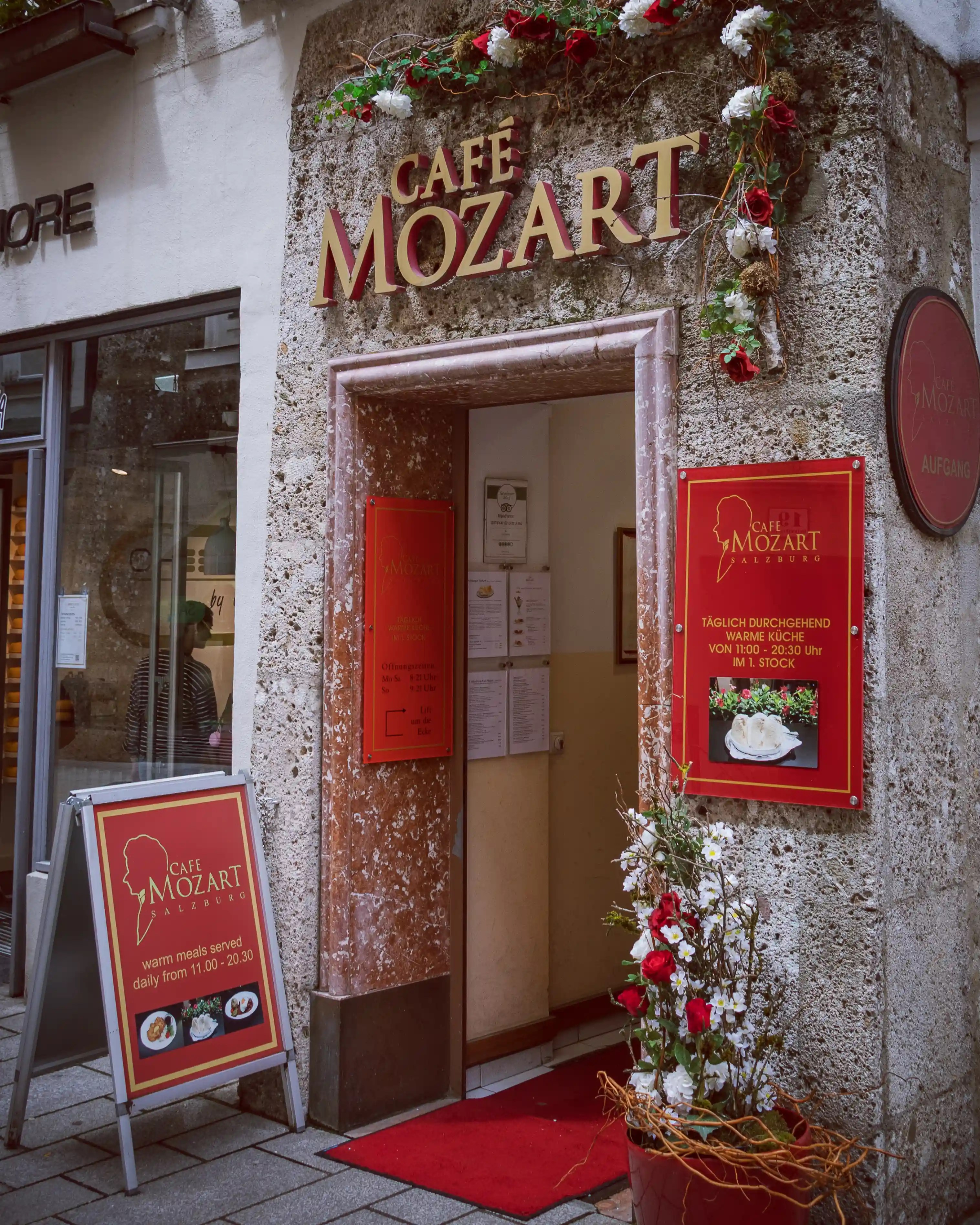The entrance to Café Mozart in Salzburg with red signage, floral decorations, and menu boards beside the doorway.