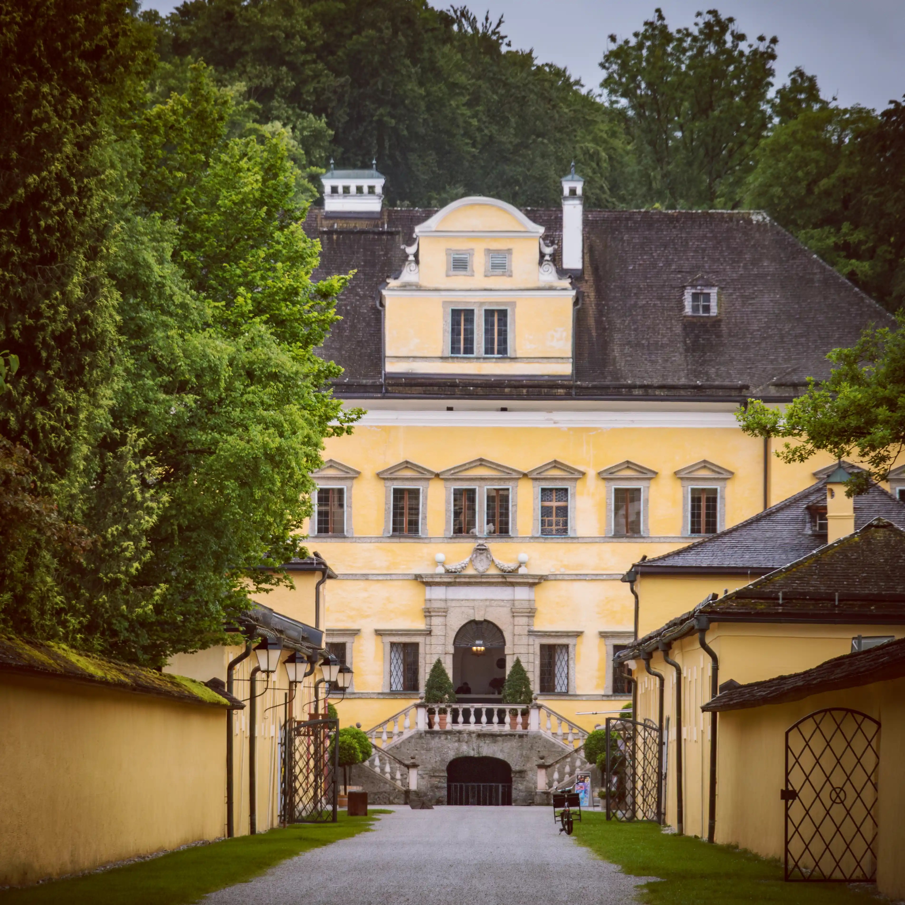 A straight gravel path leads toward the yellow façade of Hellbrunn Palace, framed by low buildings, green lawns, and trees.