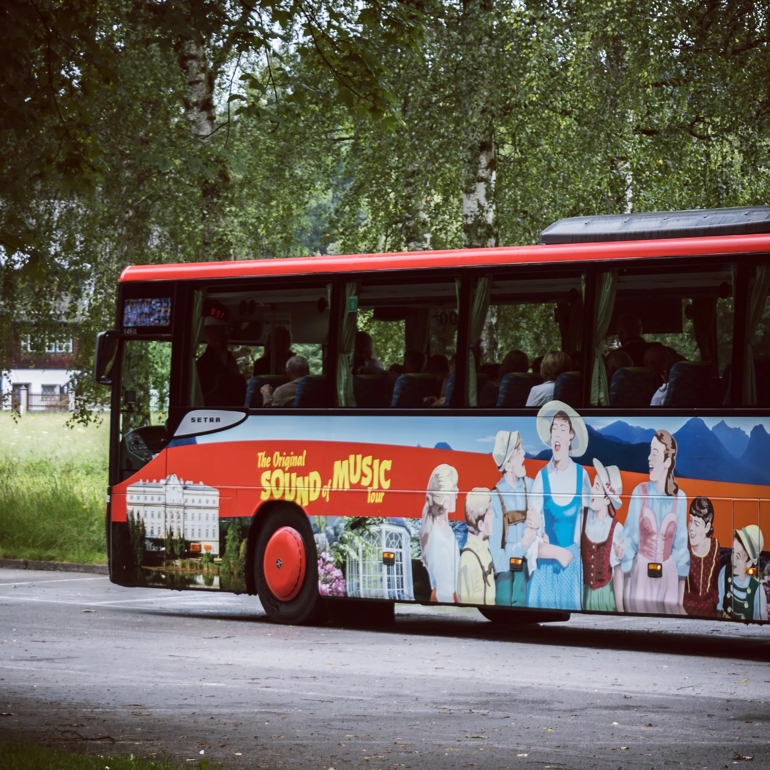 A red tour bus decorated with illustrated scenes from The Sound of Music is parked along a tree-lined road.