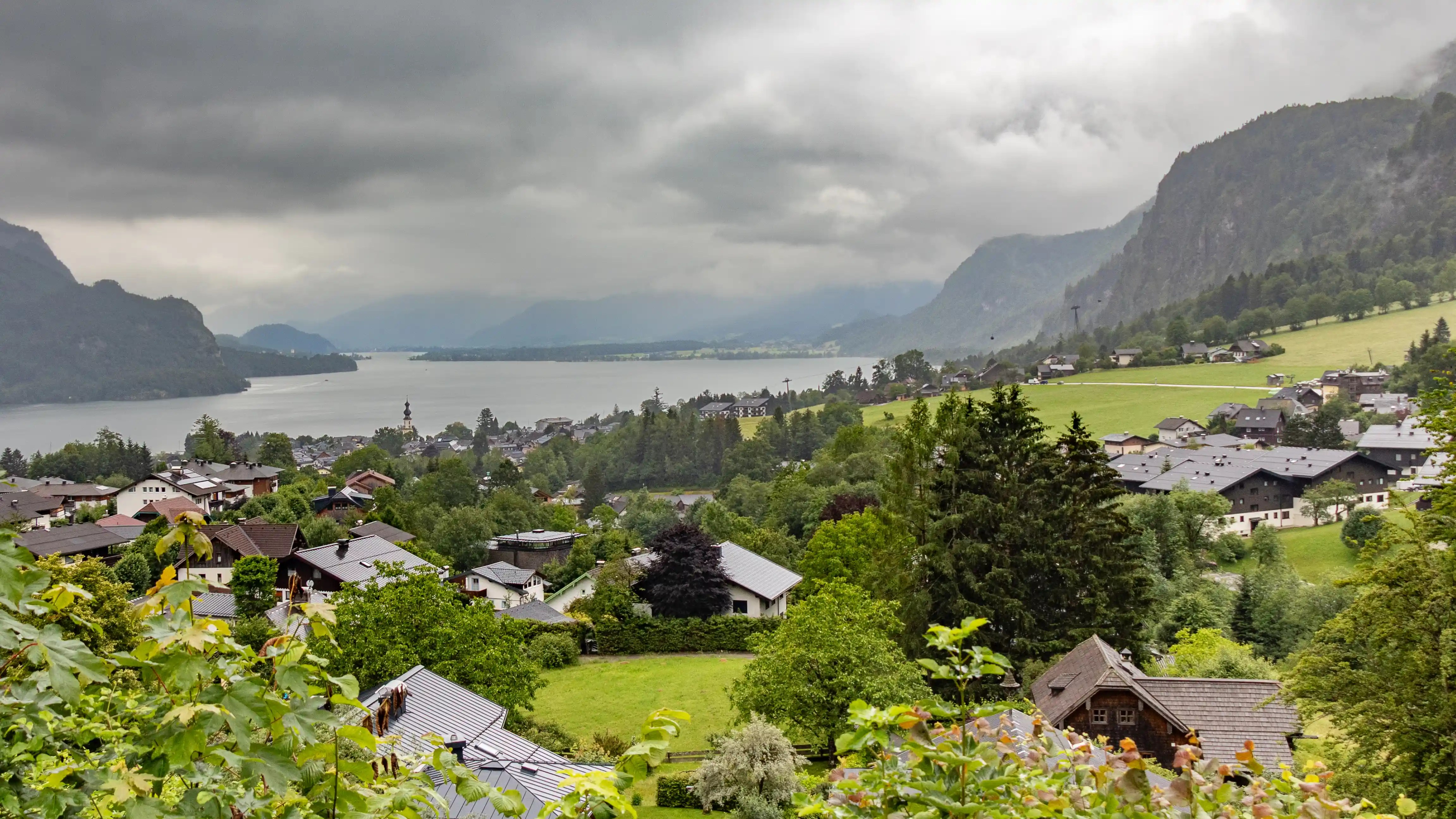 A lakeside village and rolling green hills surround a wide lake beneath low, cloudy skies.