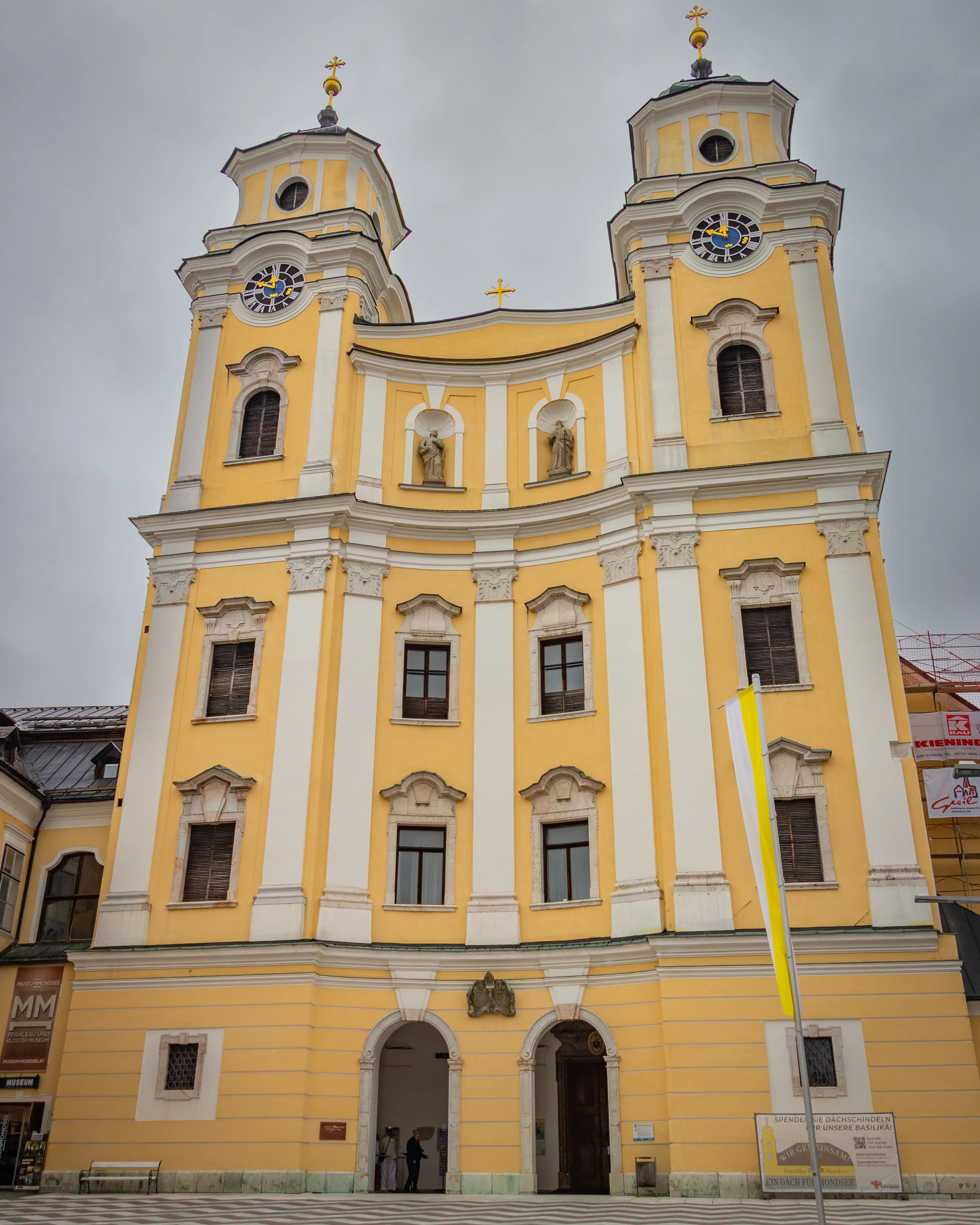 The bright yellow twin-towered façade of Basilika Mondsee rises against a cloudy sky in the town square of Mondsee.