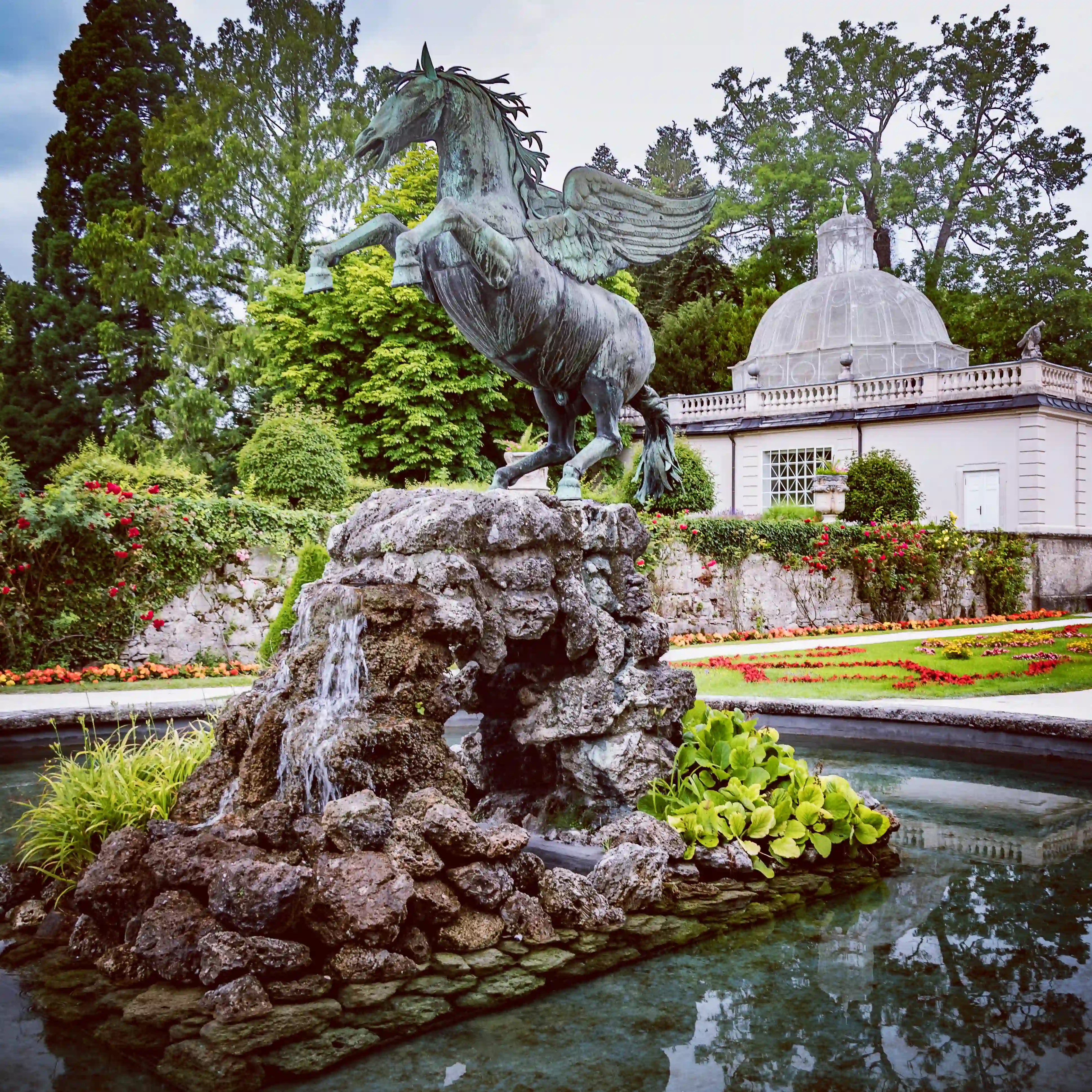 A bronze Pegasus statue rears up on a rocky fountain base surrounded by flowers and garden paths at Mirabell Gardens in Salzburg.