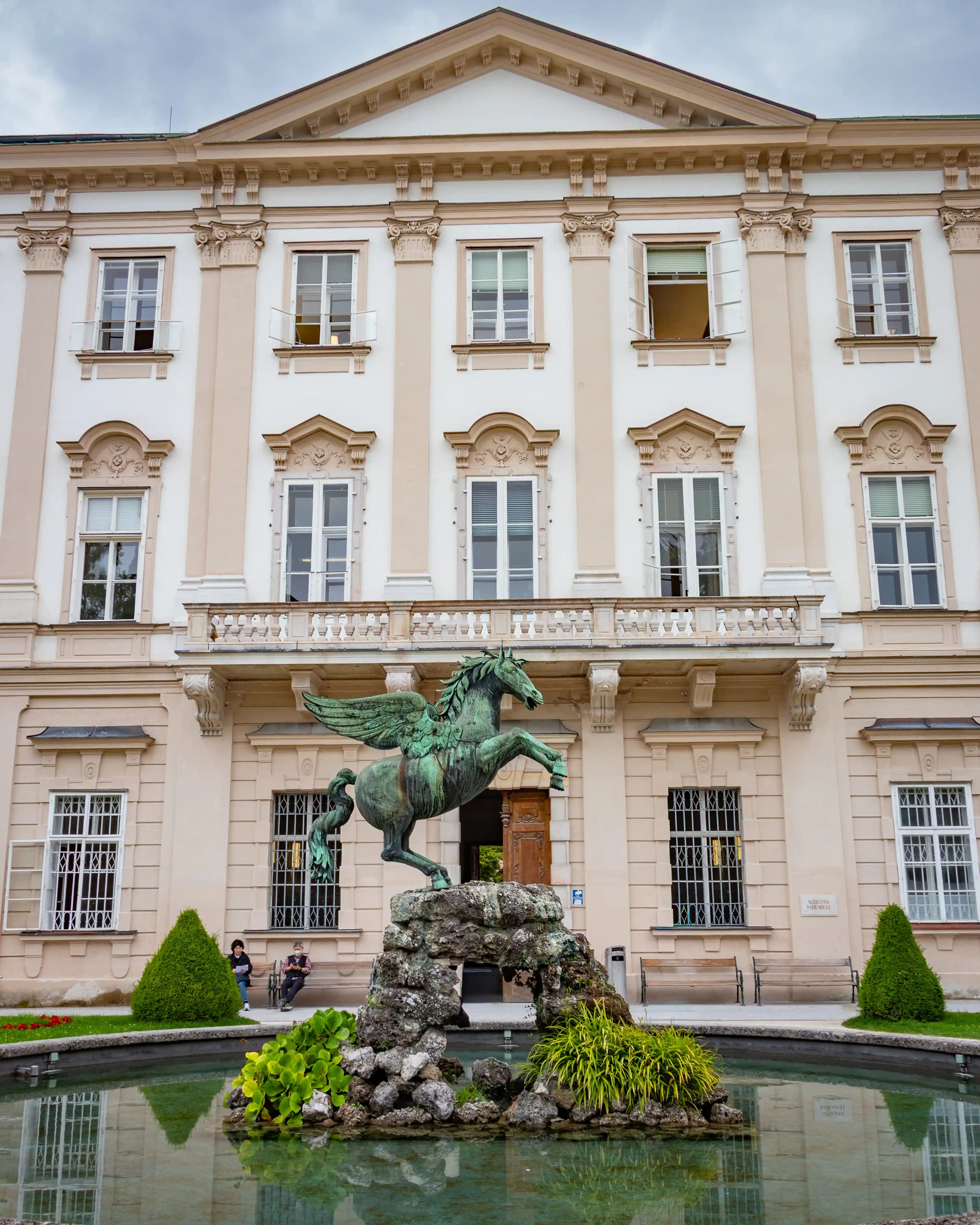 The Pegasus Fountain stands in front of the pale facade of Mirabell Palace, reflected in the still water of the fountain basin.