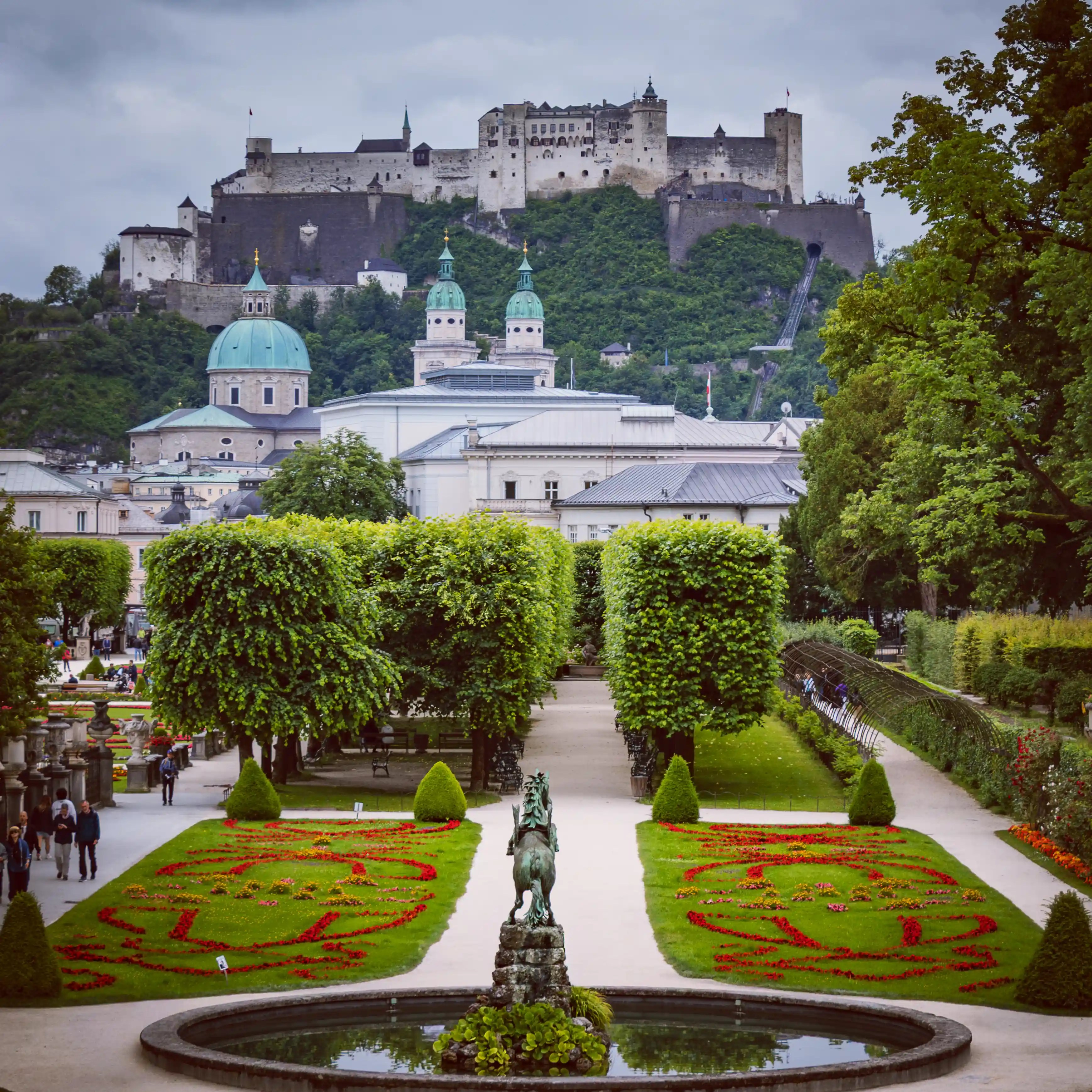 A formal garden path lined with flowerbeds and trimmed trees leads toward Hohensalzburg Fortress rising above Salzburg in the distance.