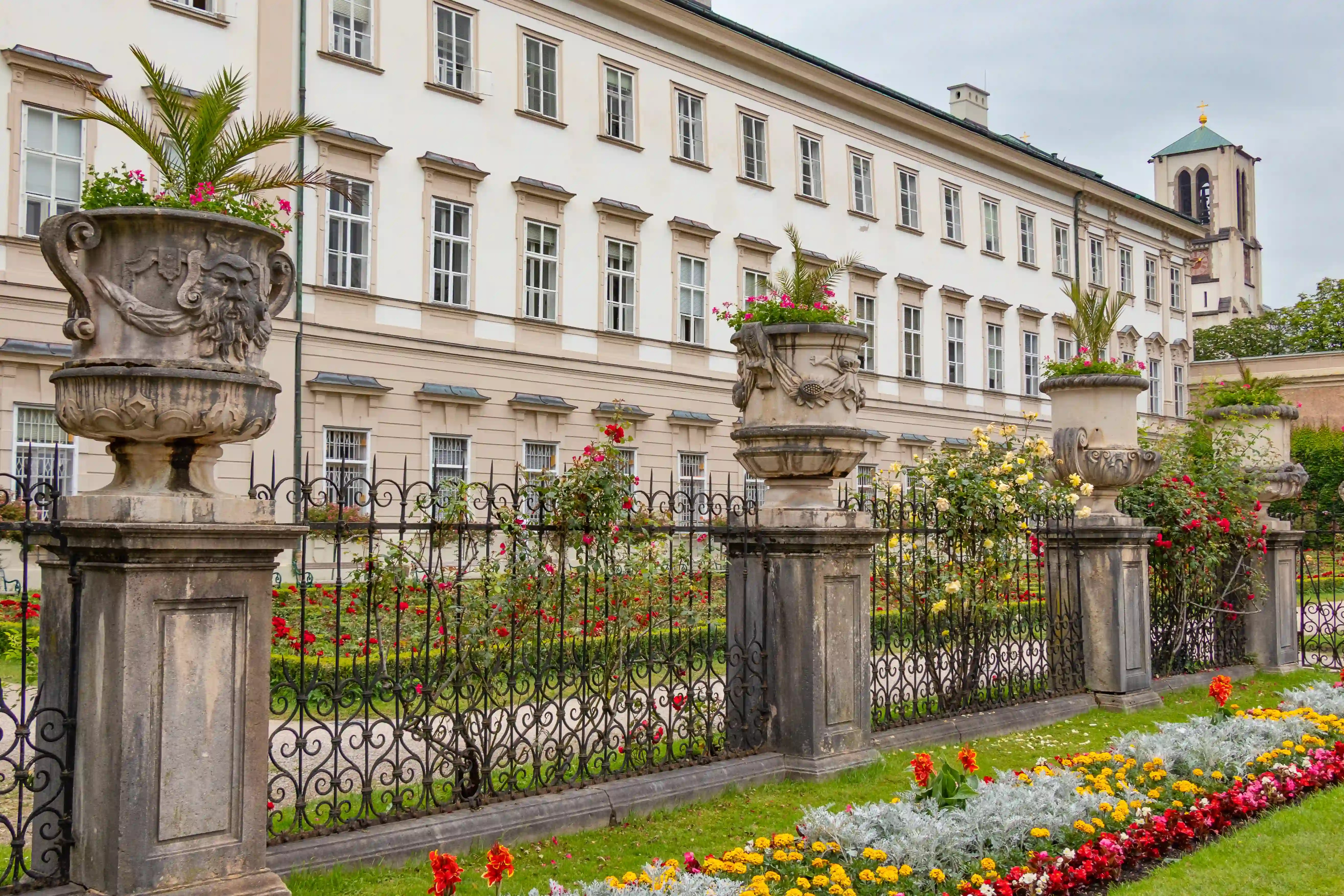 Stone urns filled with flowers line a wrought-iron fence beside colorful garden beds at Mirabell Palace.